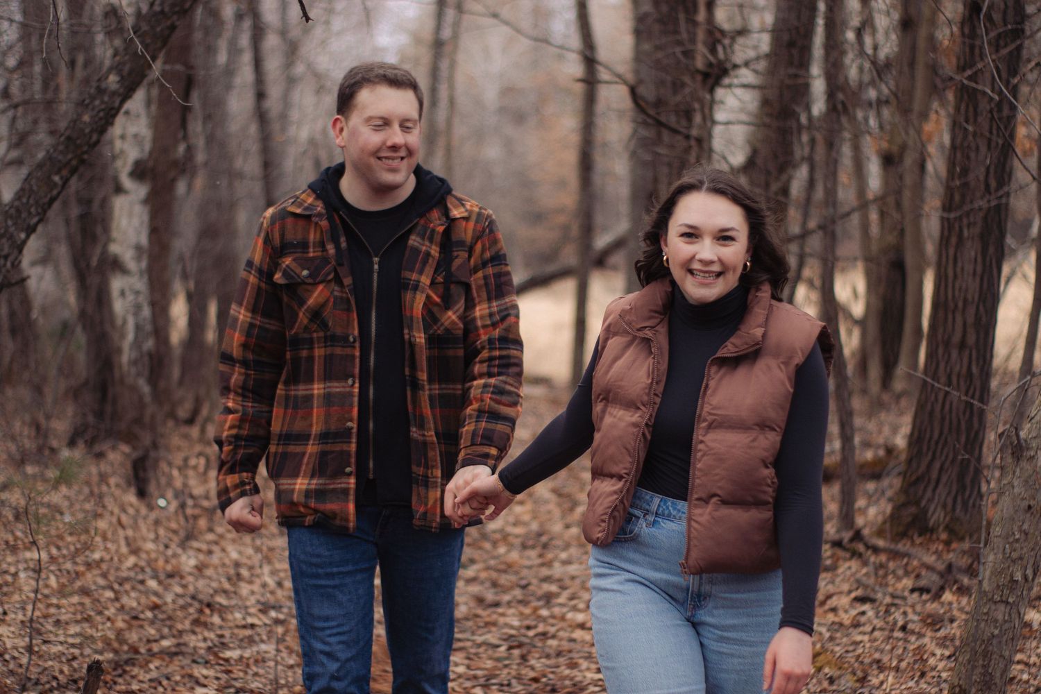 A couple walks through a wooded area during autumn wearing casual attire and plaid flannel shirts.