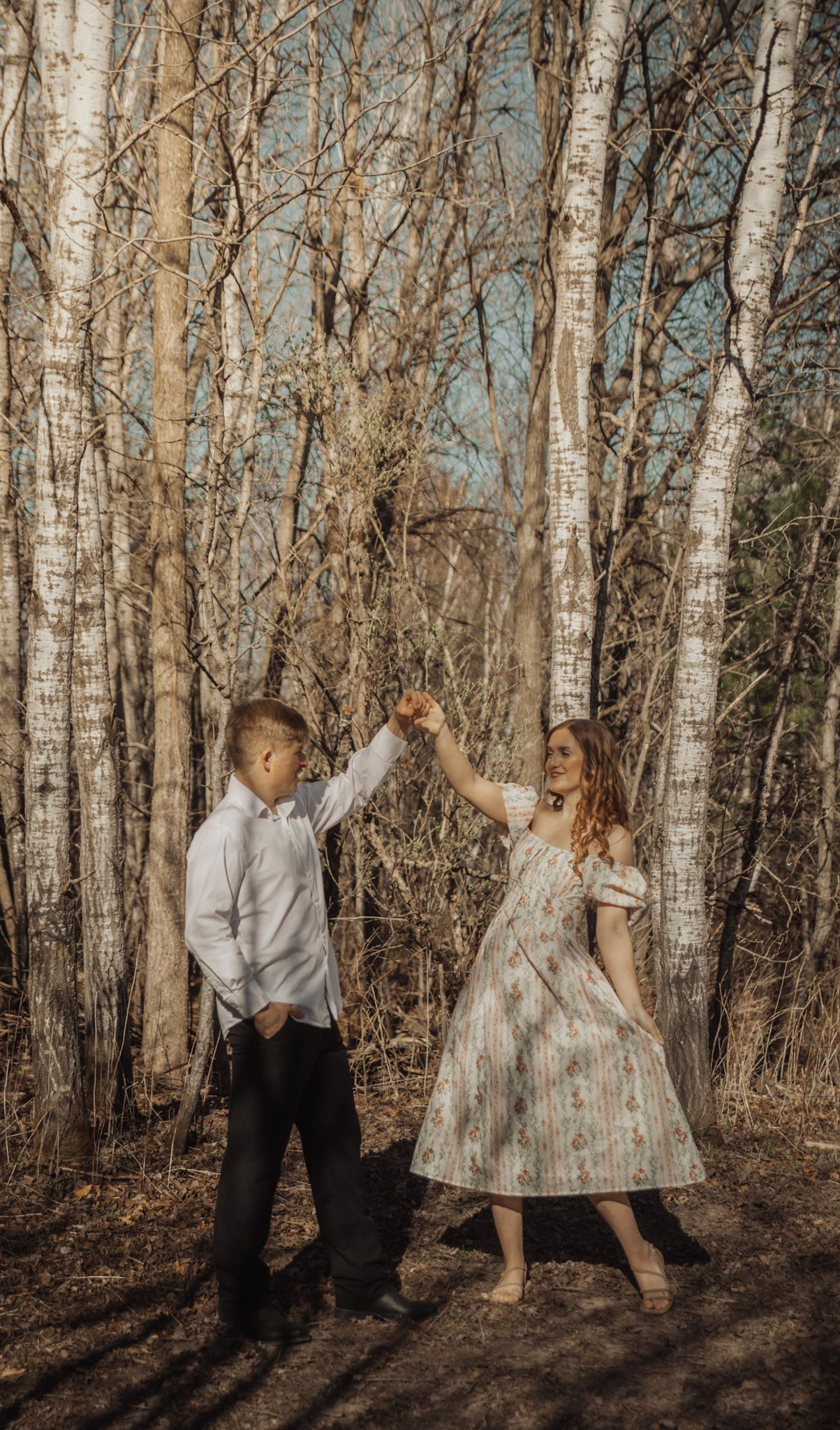 Couple dancing together in a birch forest, woman in floral dress twirling with man in white shirt.