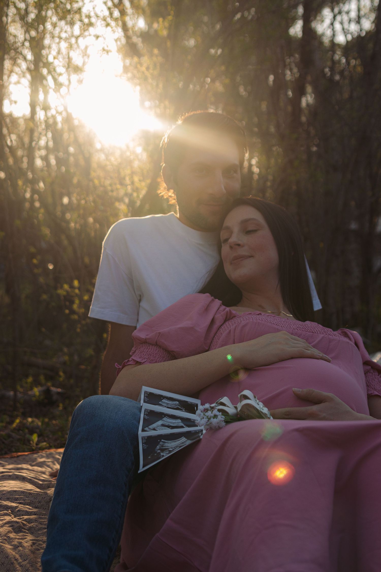A couple shares a tender moment at sunset while displaying ultrasound images in a wooded setting.