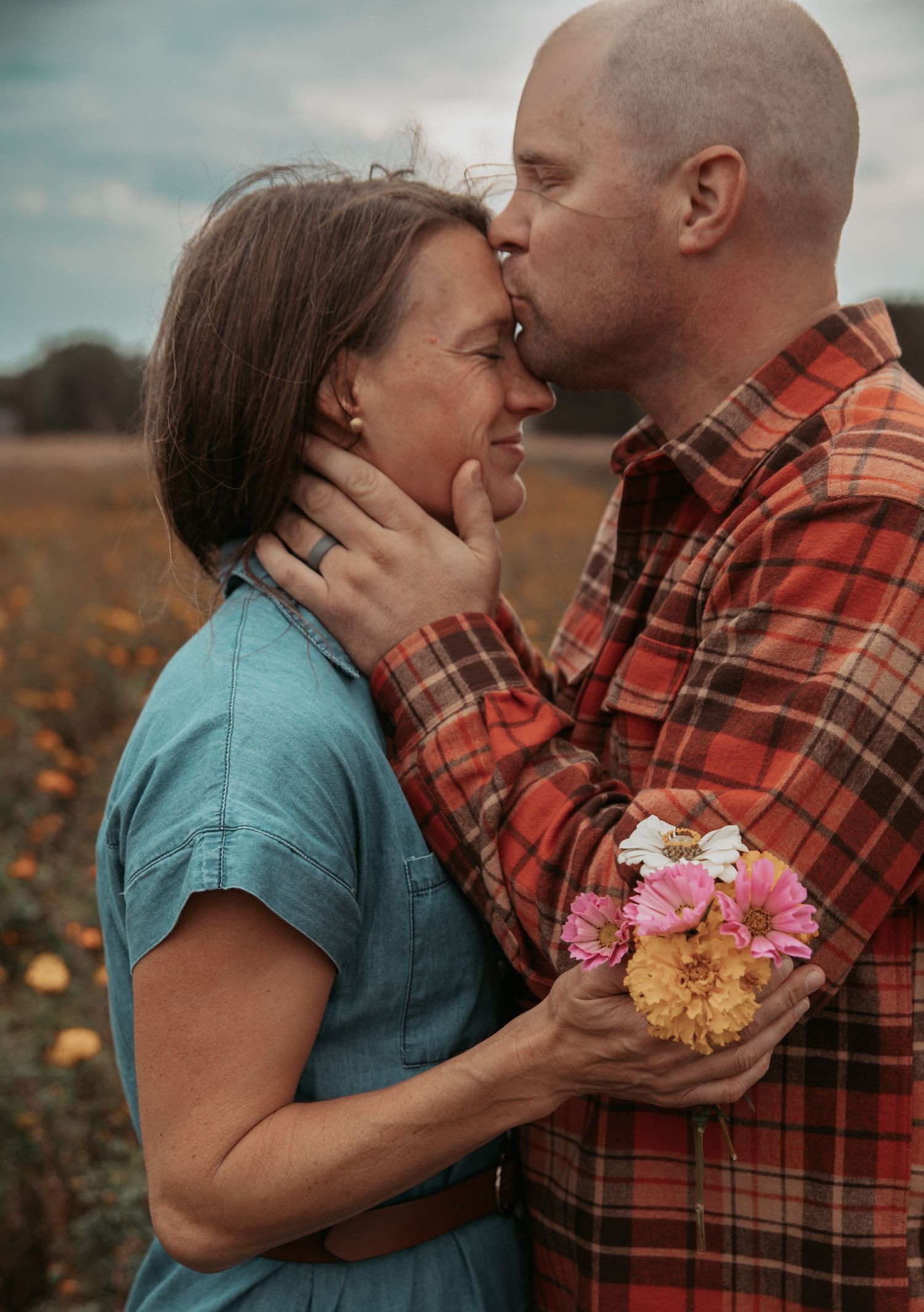 A couple shares an intimate moment in a field while holding flowers against a moody sky background.