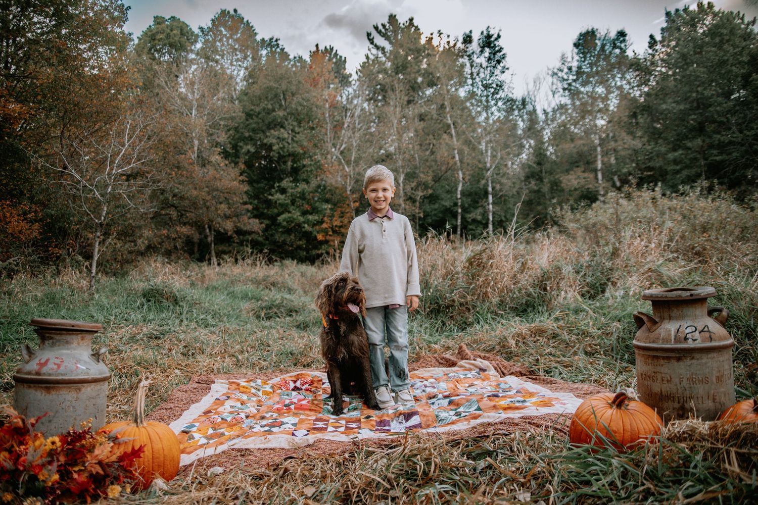 A fall themed picnic setup in a forest clearing with pumpkins and a colorful patterned blanket on the ground.