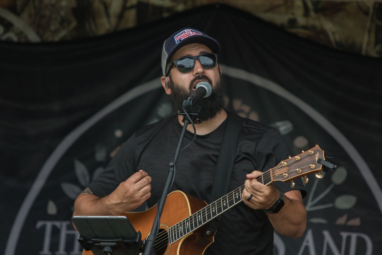 Musician performs on acoustic guitar while singing into microphone on dark stage with circular backdrop.