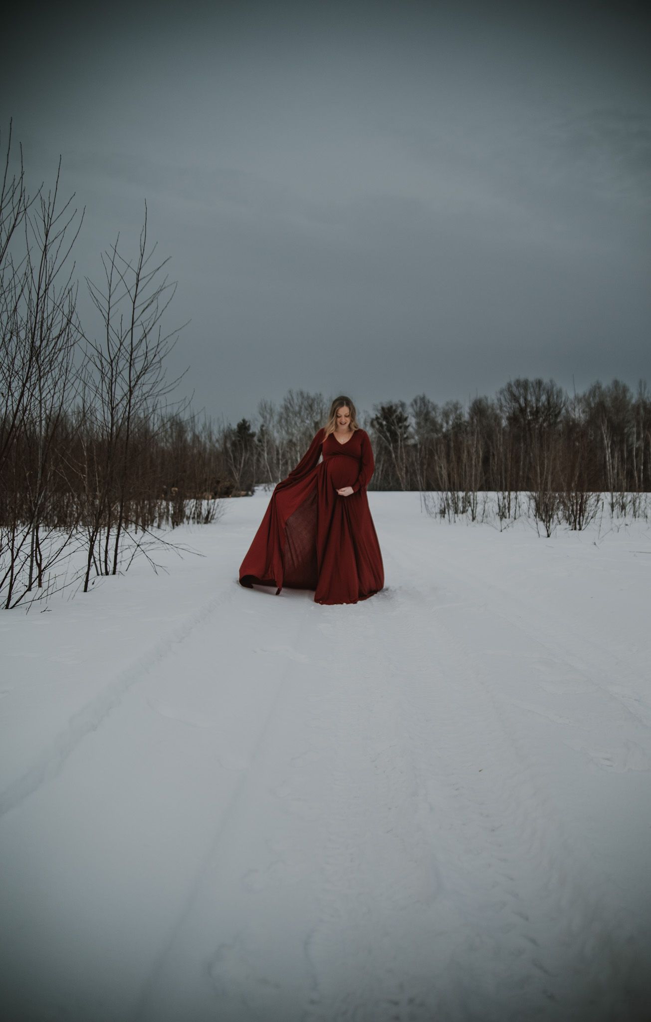 Two figures in burgundy dresses stand in distance on snow-covered field with bare trees.
