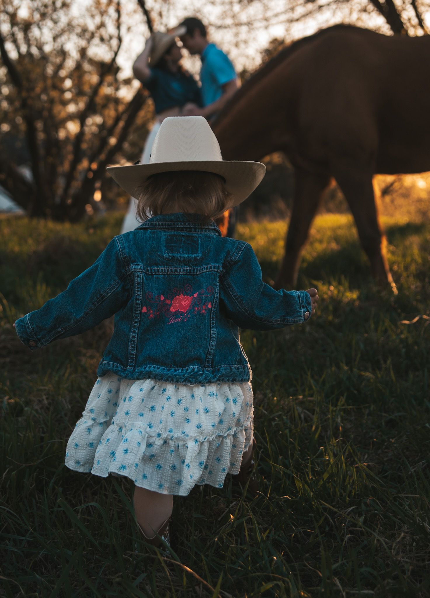 A toddler in a denim jacket, floral dress, and cowboy hat walks toward a person with a horse at golden hour.