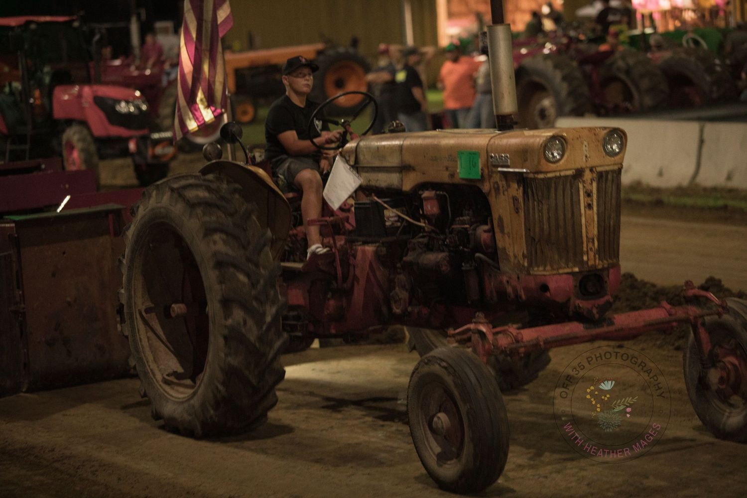 Row of vintage tractors and farm equipment displayed on dirt ground at night with warm lighting.