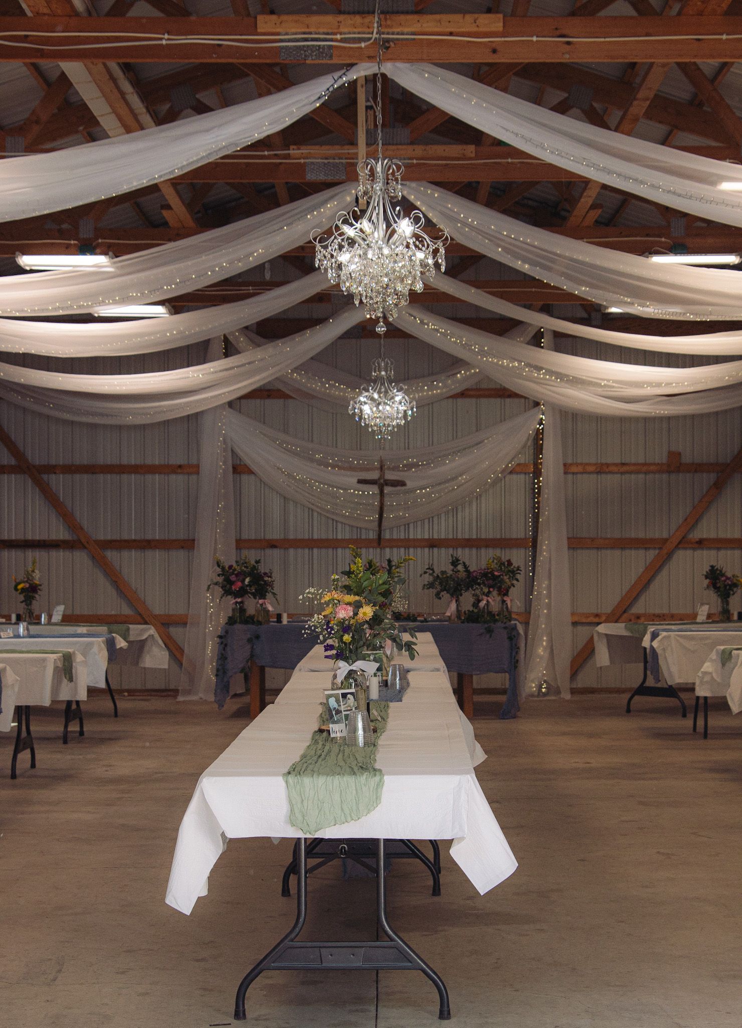 A rustic wedding reception venue with white fabric draping, crystal chandeliers, and a long table setting with floral centerpieces.