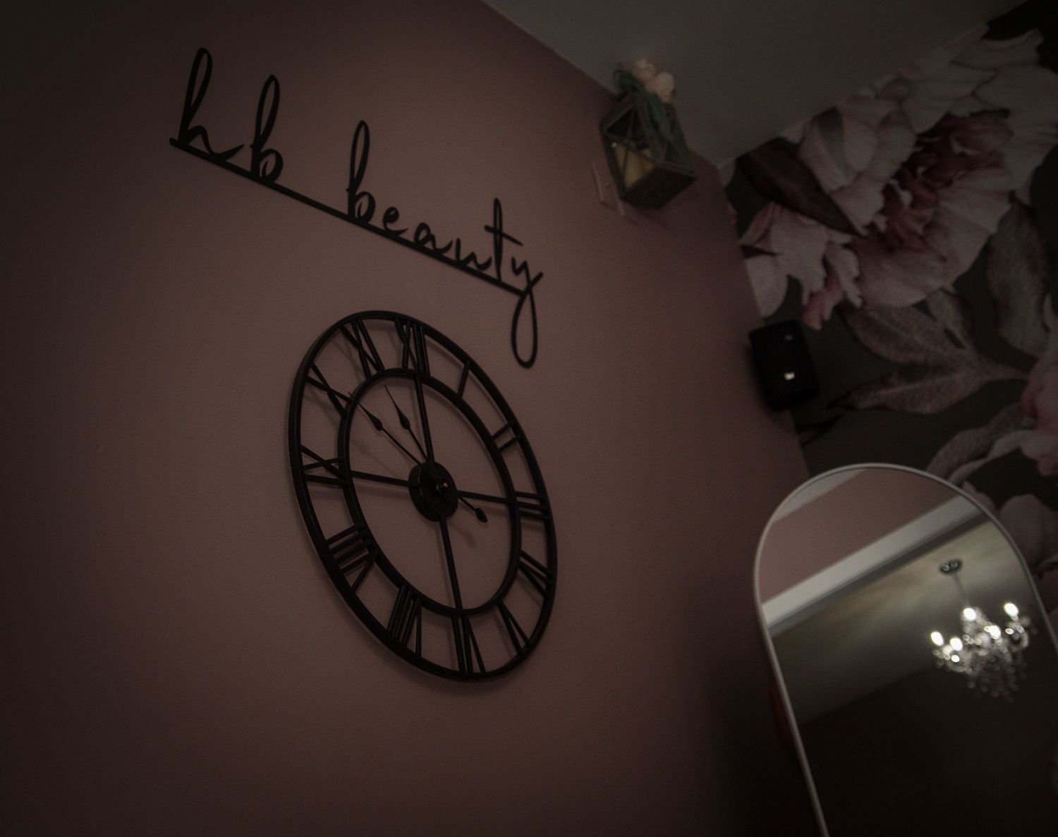 Decorative wall clock made of black metal mounted on cream colored wall beneath string lights.