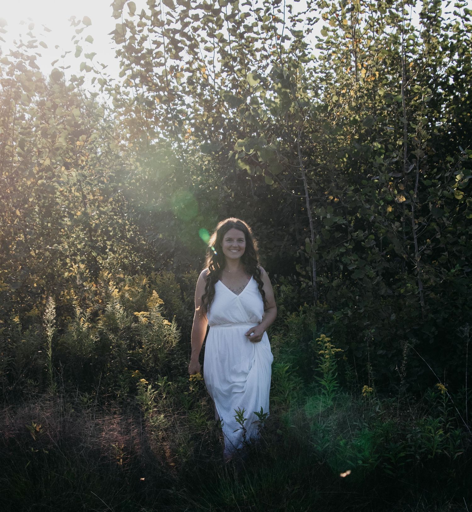 A figure stands in an outdoor setting at sunset, backlit by golden light filtering through trees in the background.