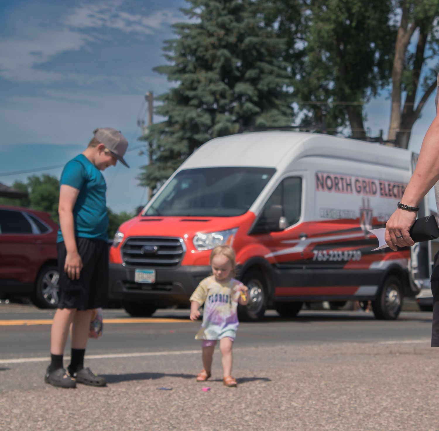 A small child stands with an adult in front of a red and white delivery van on a sunny day.