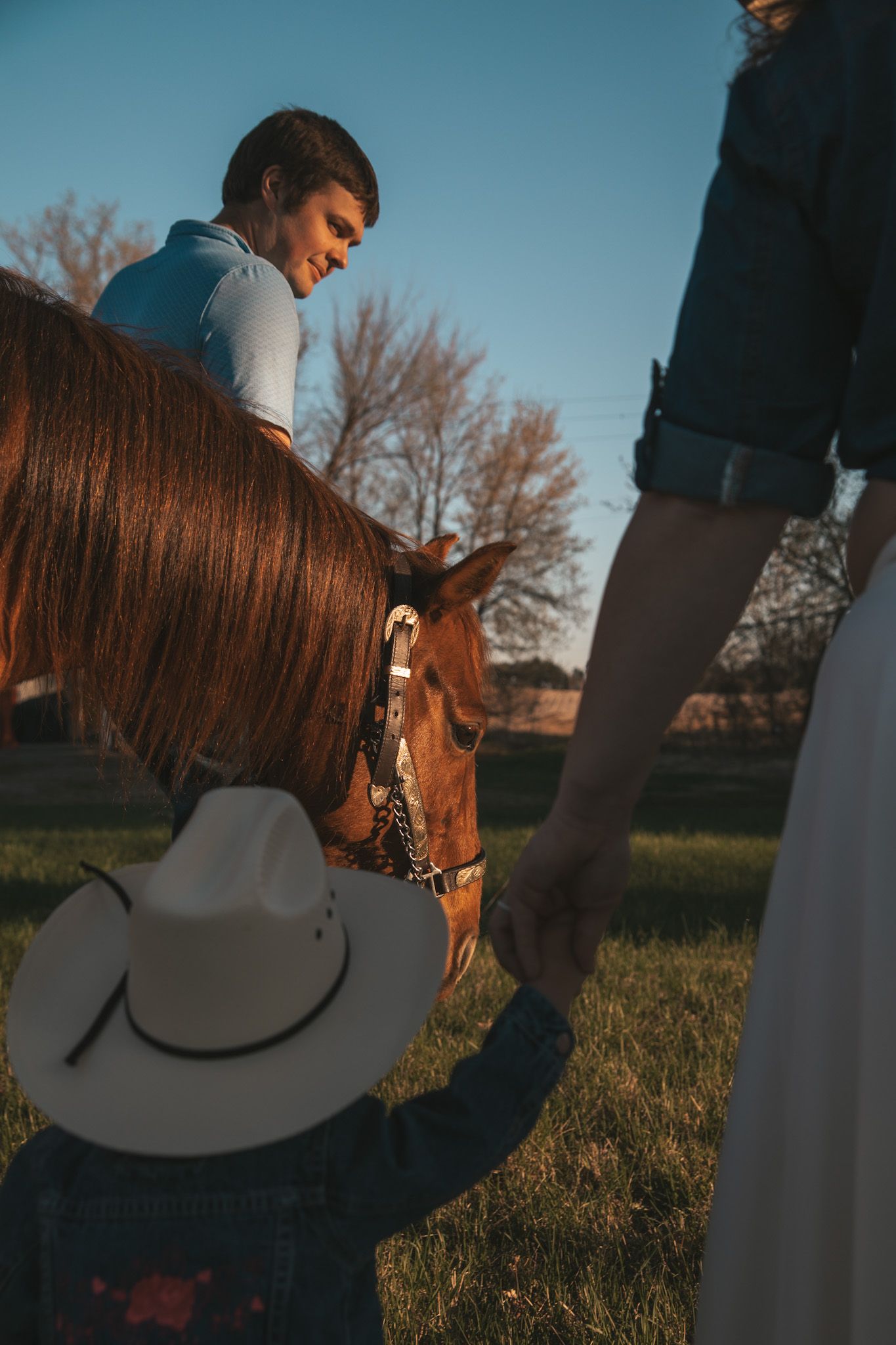Young child in white cowboy hat holds hand of pregnant woman while man on horseback smiles at sunset.