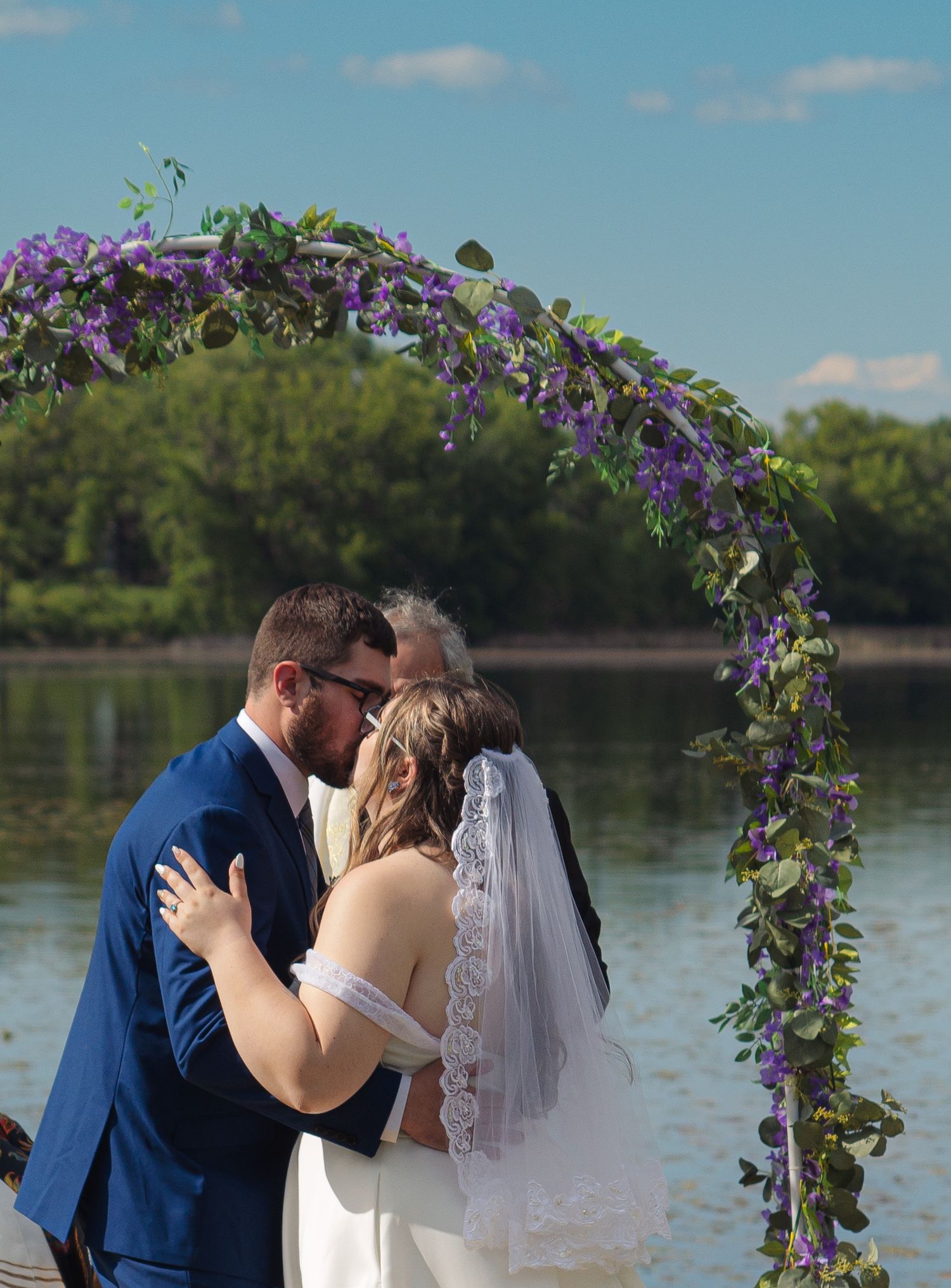 Wedding ceremony under a purple floral arch next to a serene lake on a beautiful summer day.