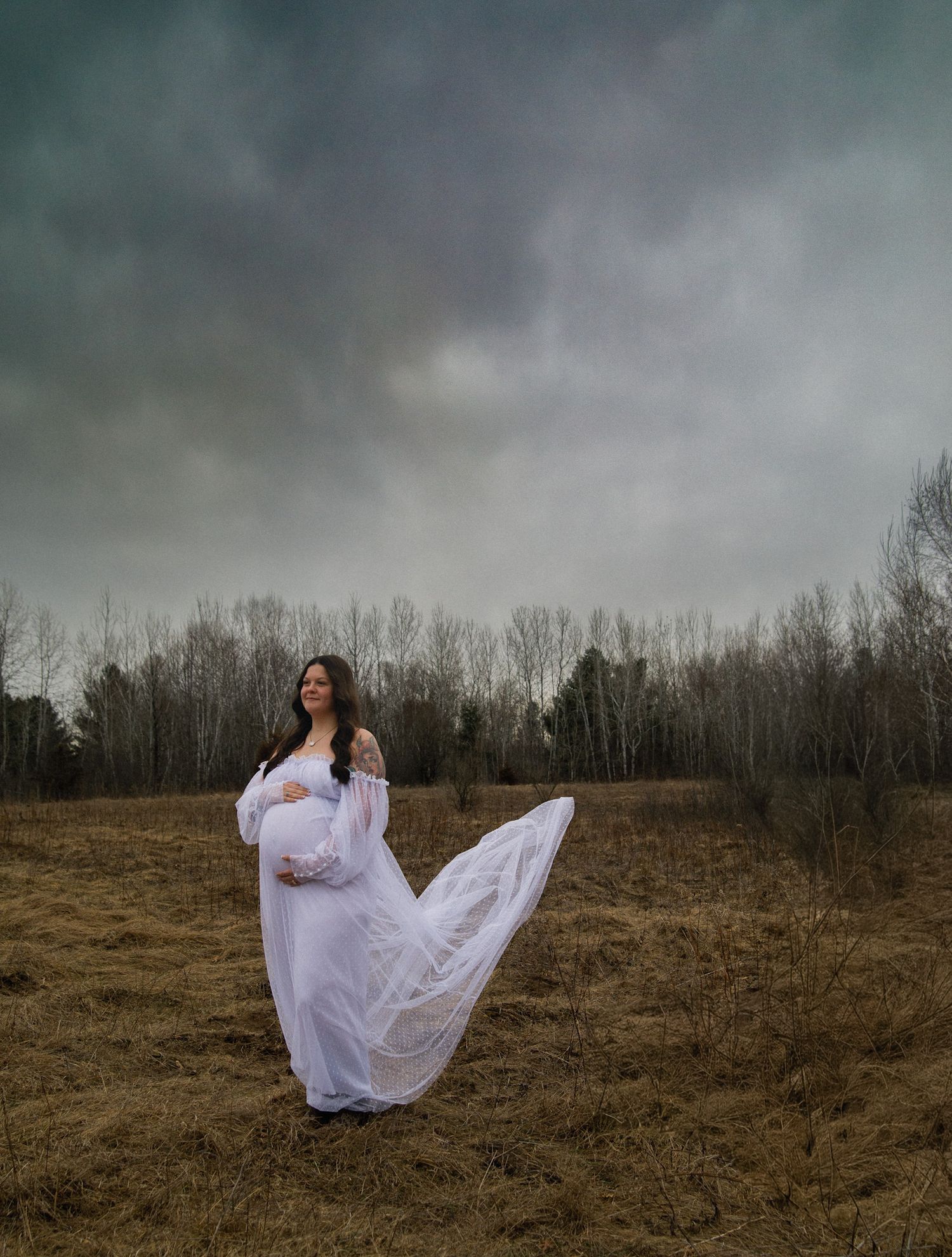 Pregnant woman in flowing white gown standing in a field under dramatic cloudy skies.