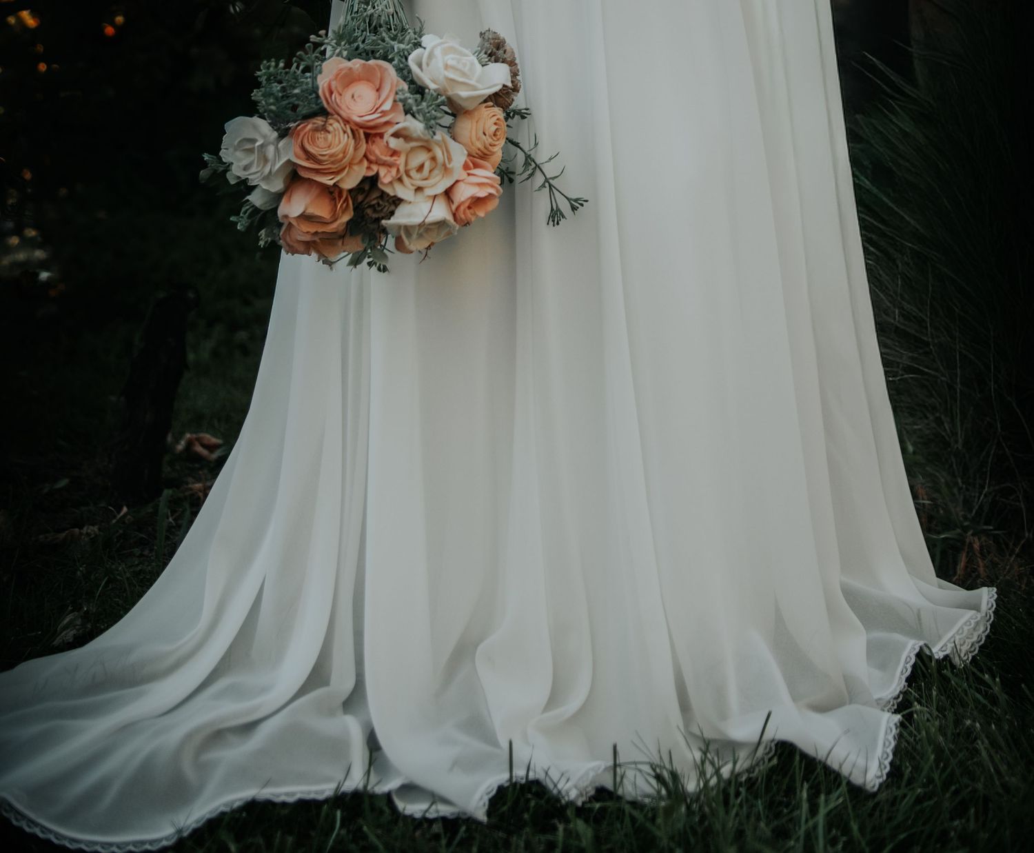 A close-up view of a flowing white wedding dress with delicate peach roses against a dark background.