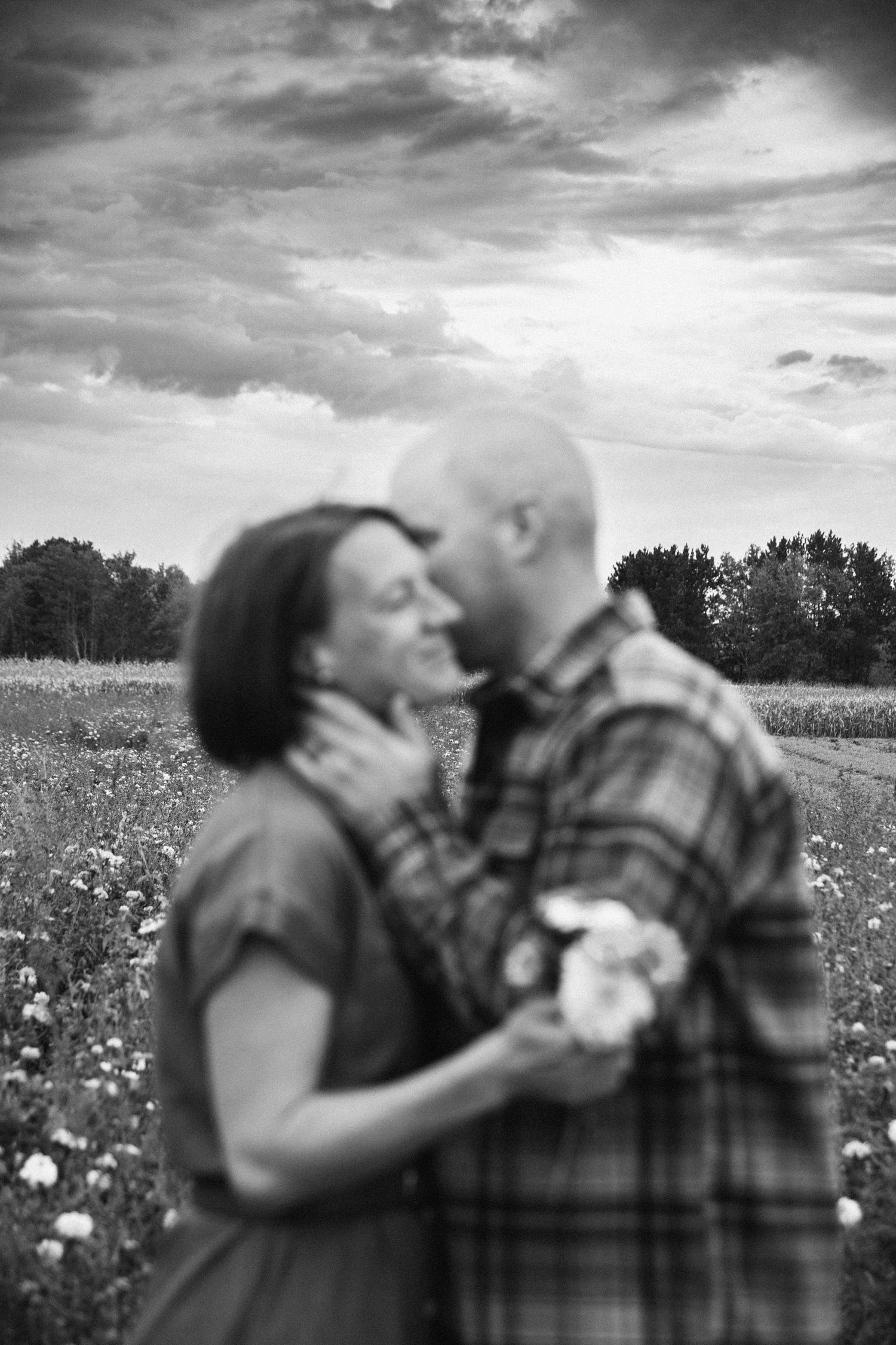 A romantic black and white portrait of a couple embracing in a wildflower meadow under dramatic cloudy skies.