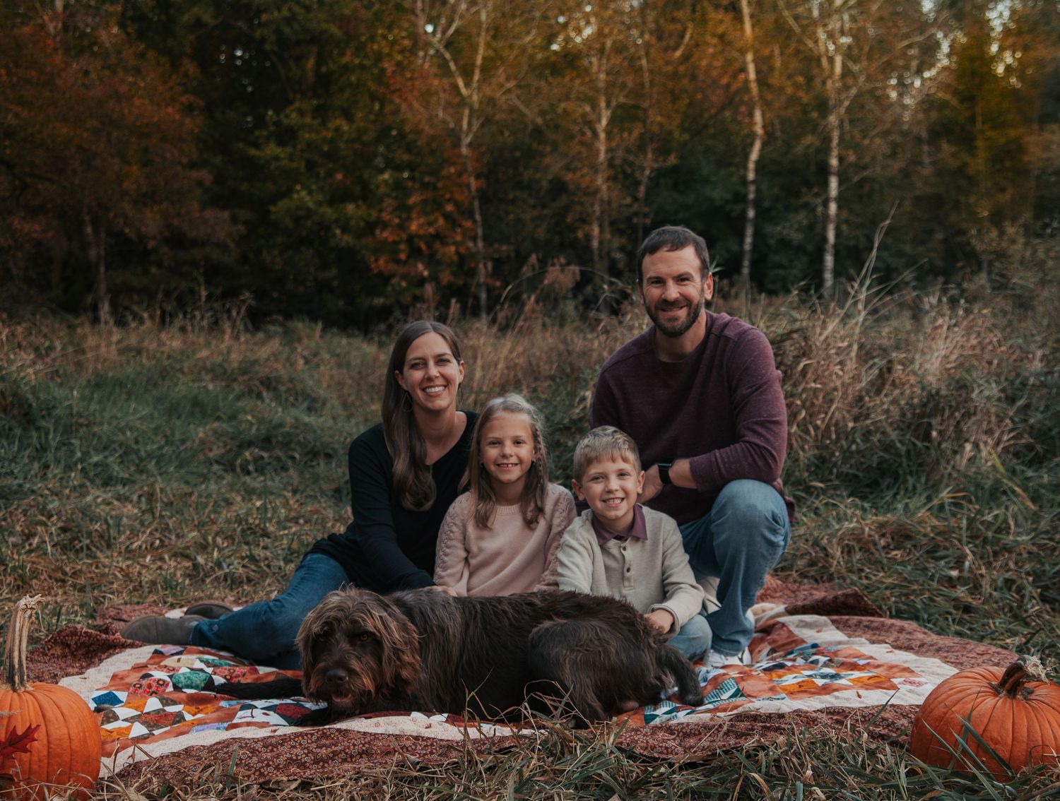 A family sits together on a cozy autumn picnic blanket surrounded by pumpkins and fall foliage.