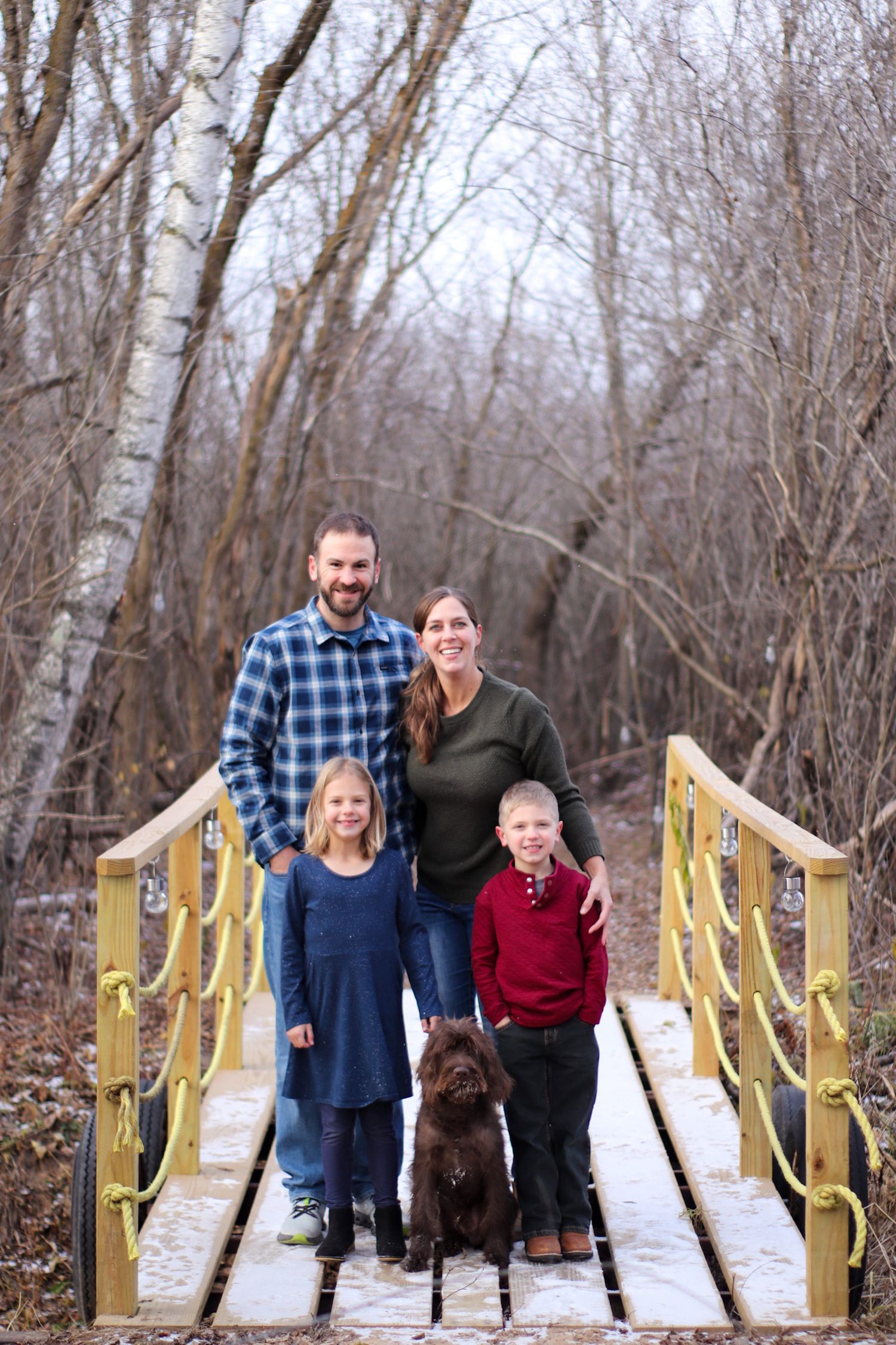 A group poses with their chocolate Labrador dog on a yellow-railed wooden footbridge in a winter forest setting.