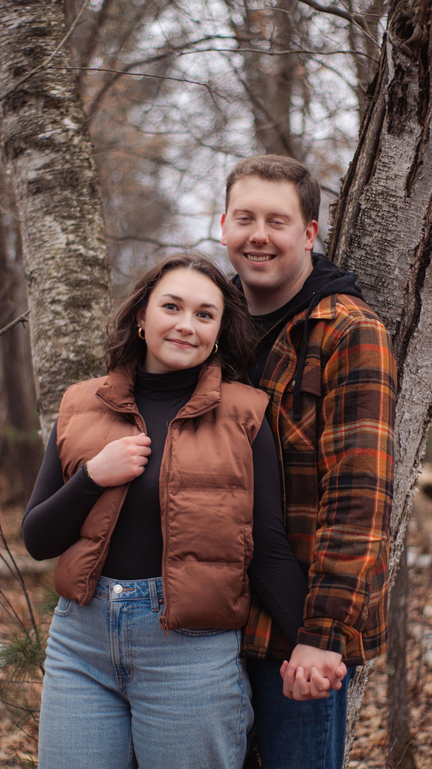 A couple in autumn clothing poses together in a forest setting with birch trees in the background.