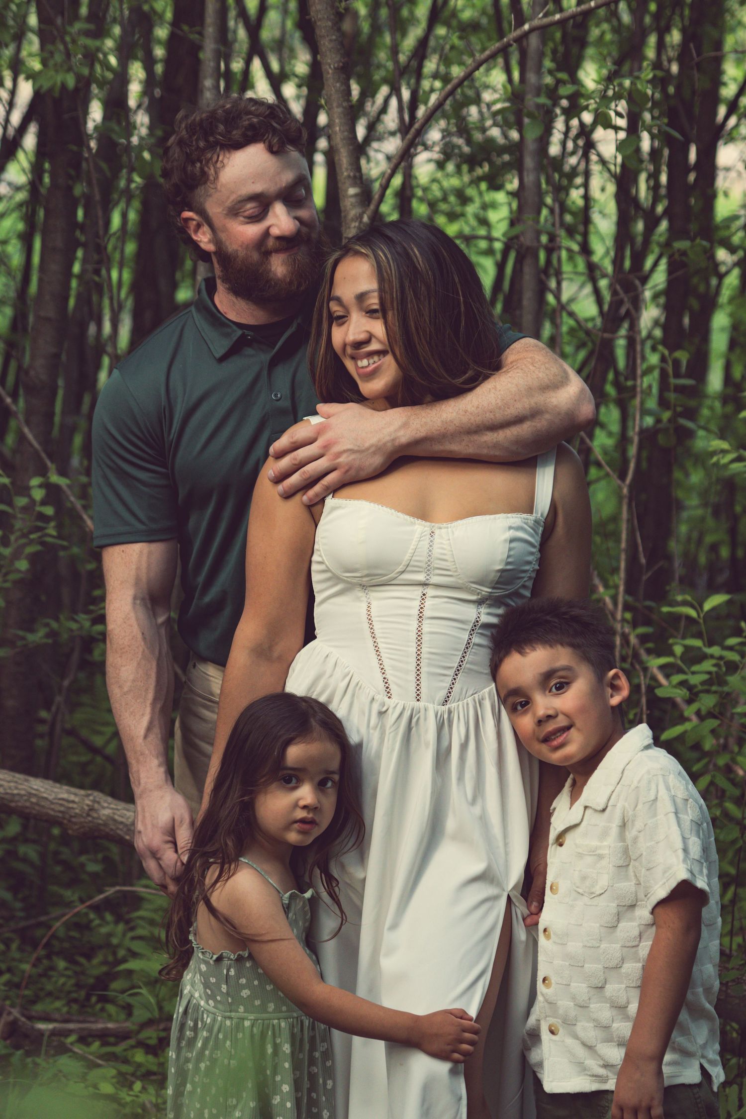 A family embraces in a peaceful forest setting, with the mother wearing a white dress and surrounded by lush greenery.