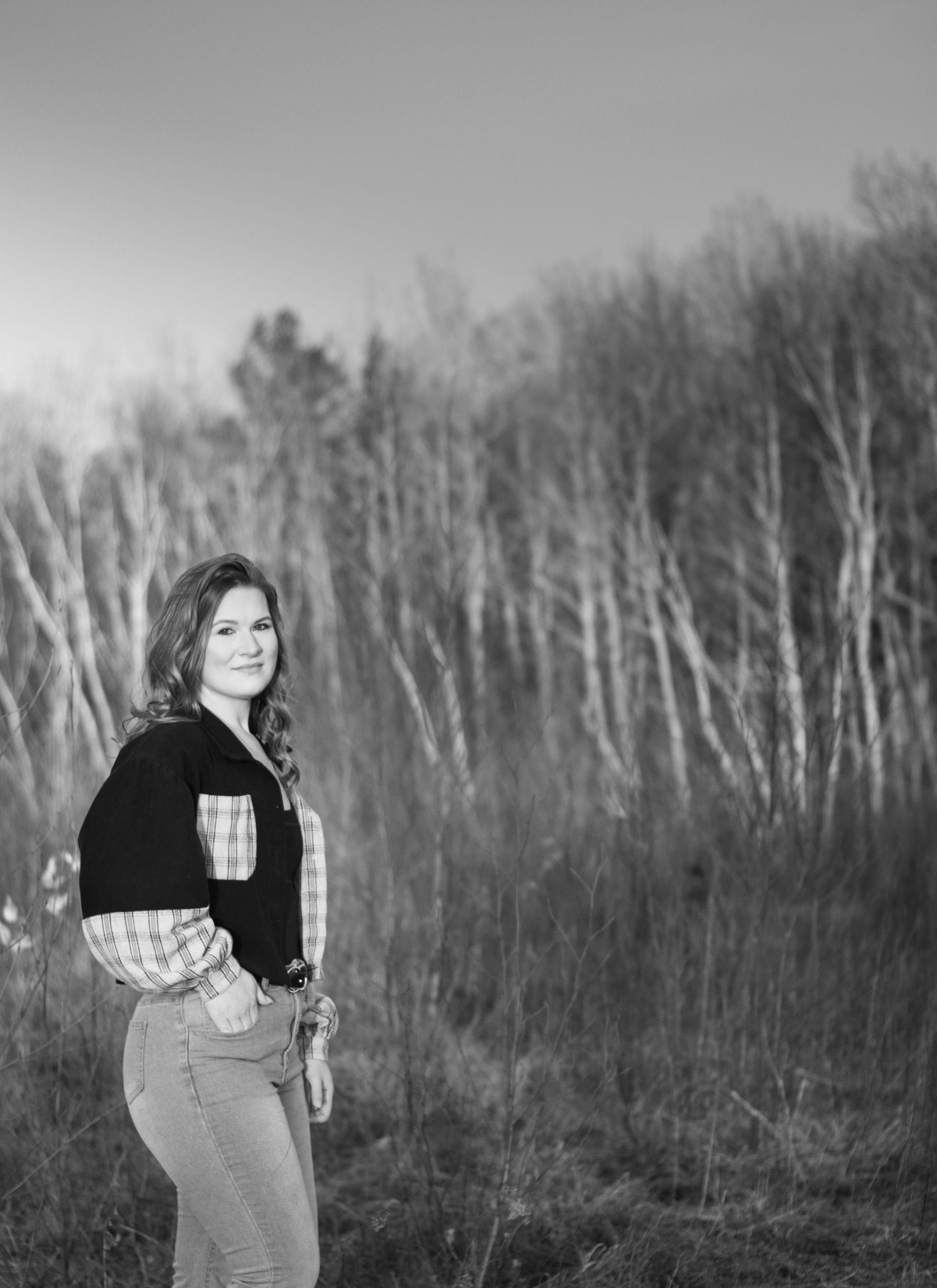 Black and white portrait of a young woman in plaid shirt standing in a field with bare trees behind her.