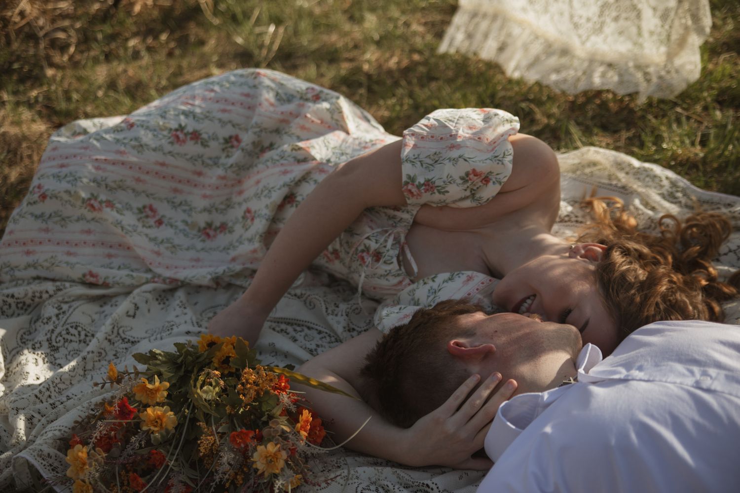 Couple lying on a floral blanket outdoors, woman in vintage dress leaning over man surrounded by wildflowers.