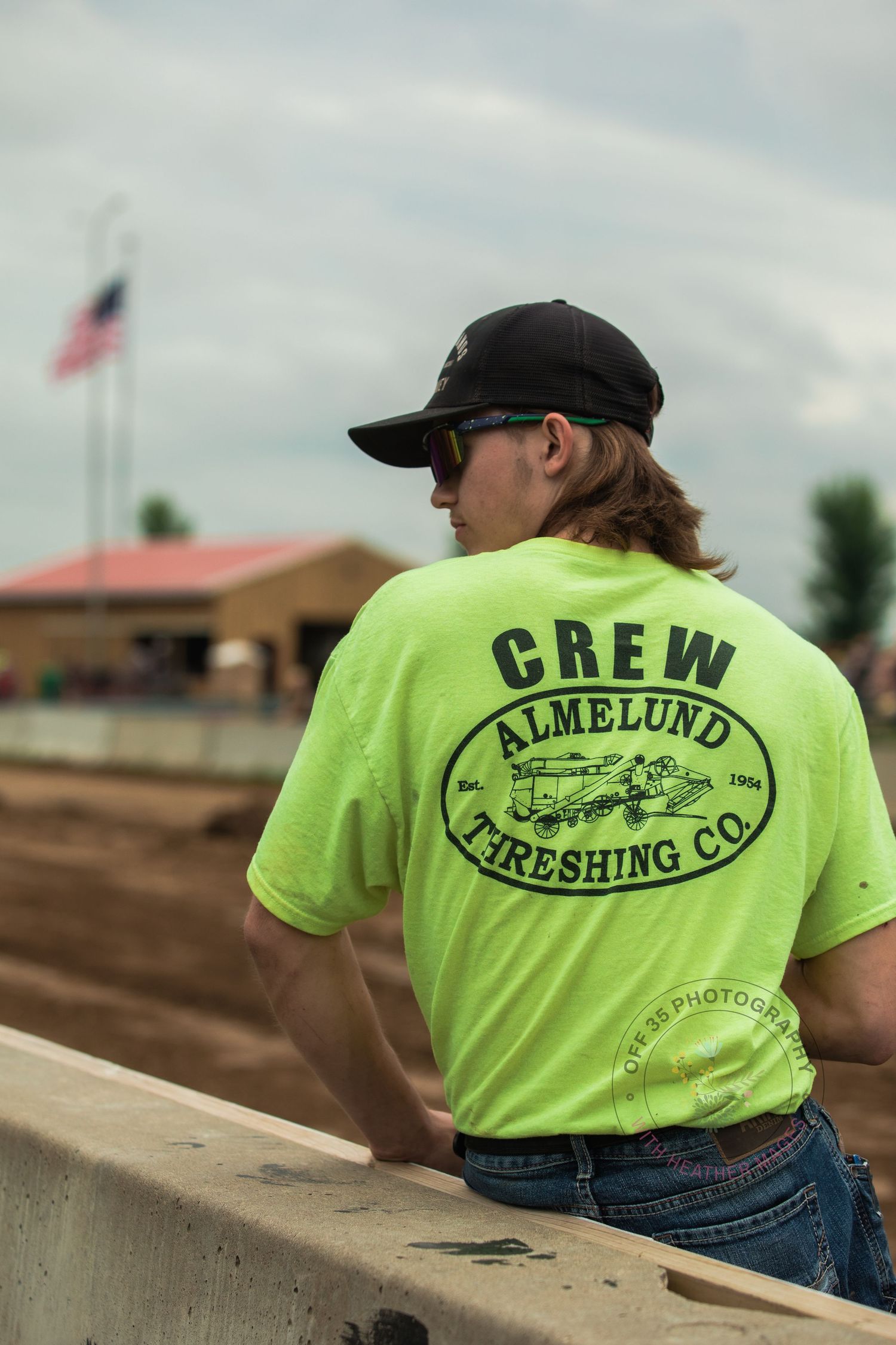 Person wearing neon yellow Crew Threshing Co t-shirt sitting on concrete ledge outdoors.
