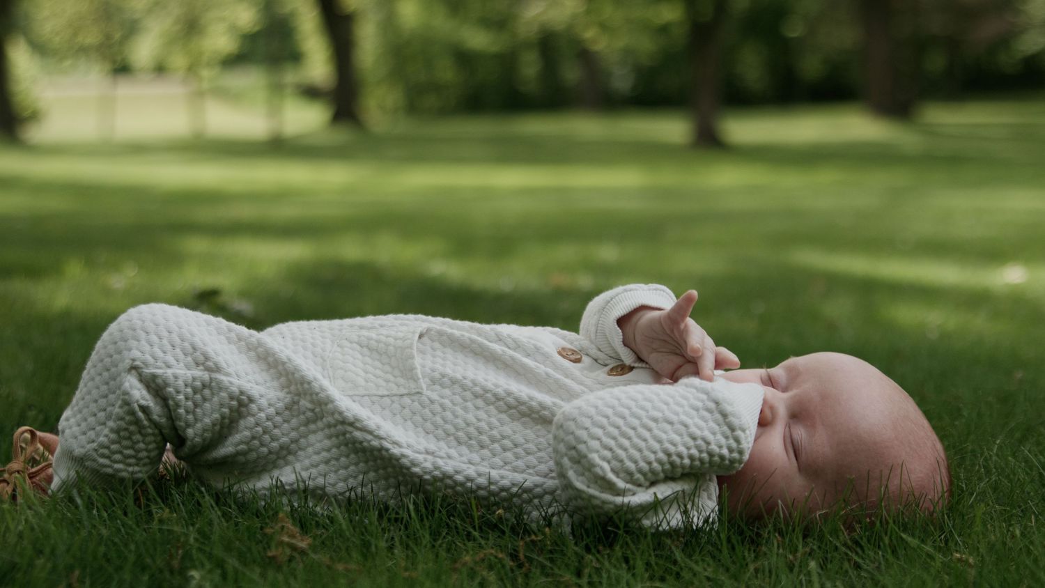 A baby lies on a green lawn wearing a white onesie while looking up at the sky on a sunny day.