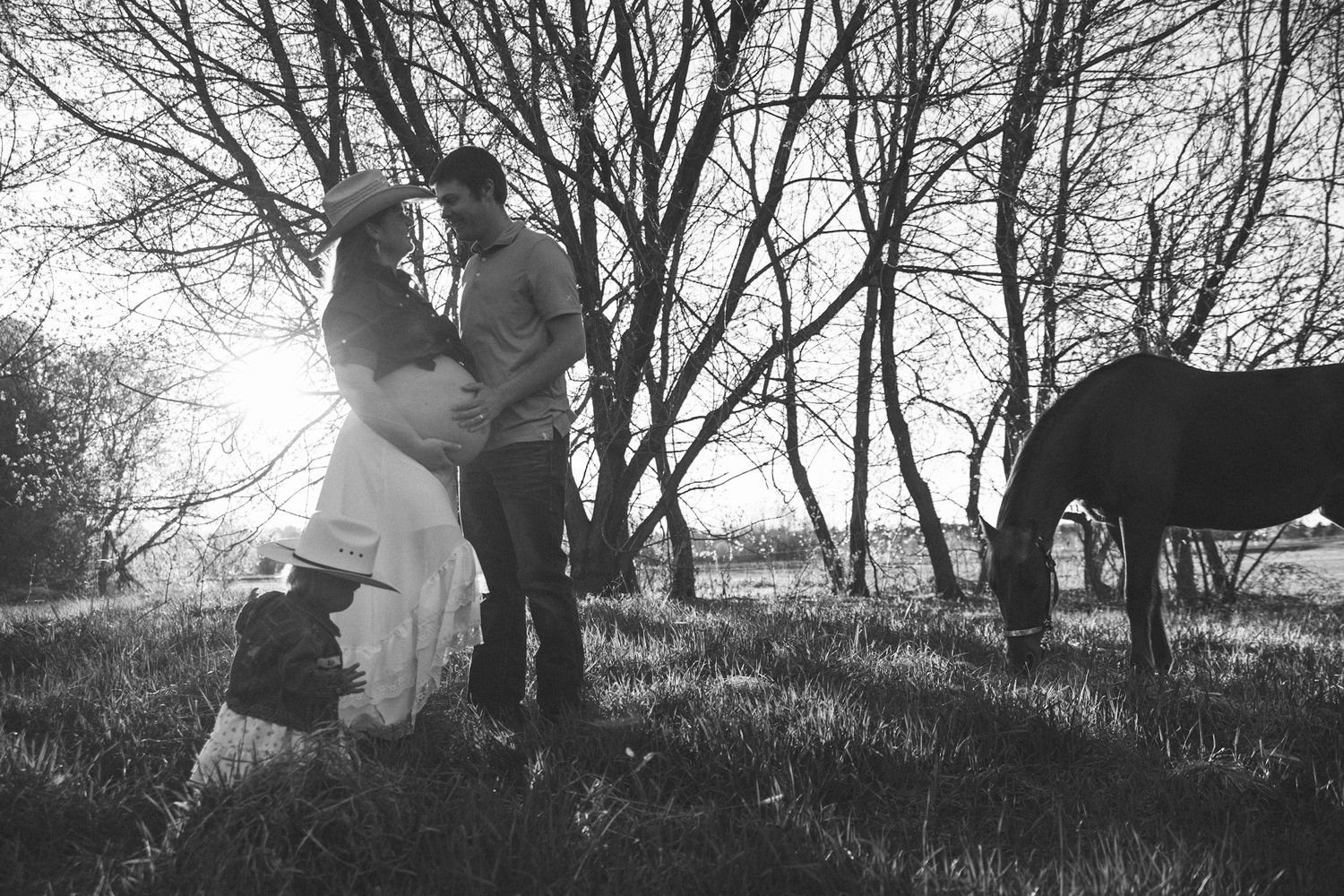 Black and white maternity photo of couple embracing near trees with a horse grazing in background.