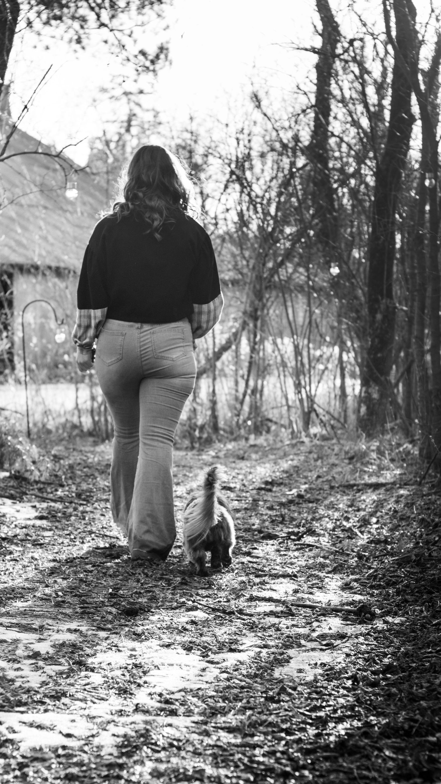 Woman in flare jeans walking a small dog on a muddy rural path, photographed from behind in black and white.