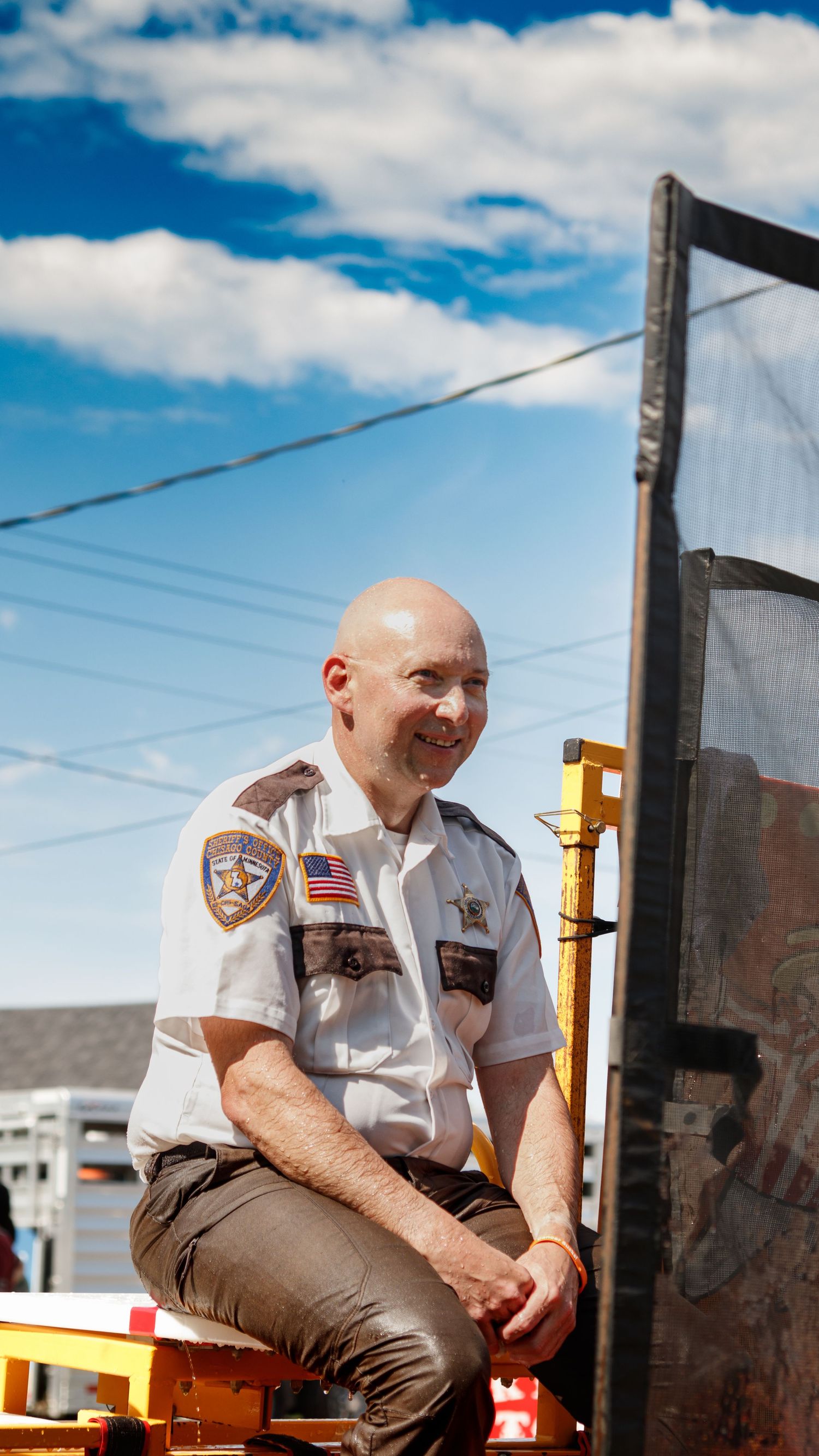 A security officer in uniform sits on metal equipment against a bright blue sky with white clouds.
