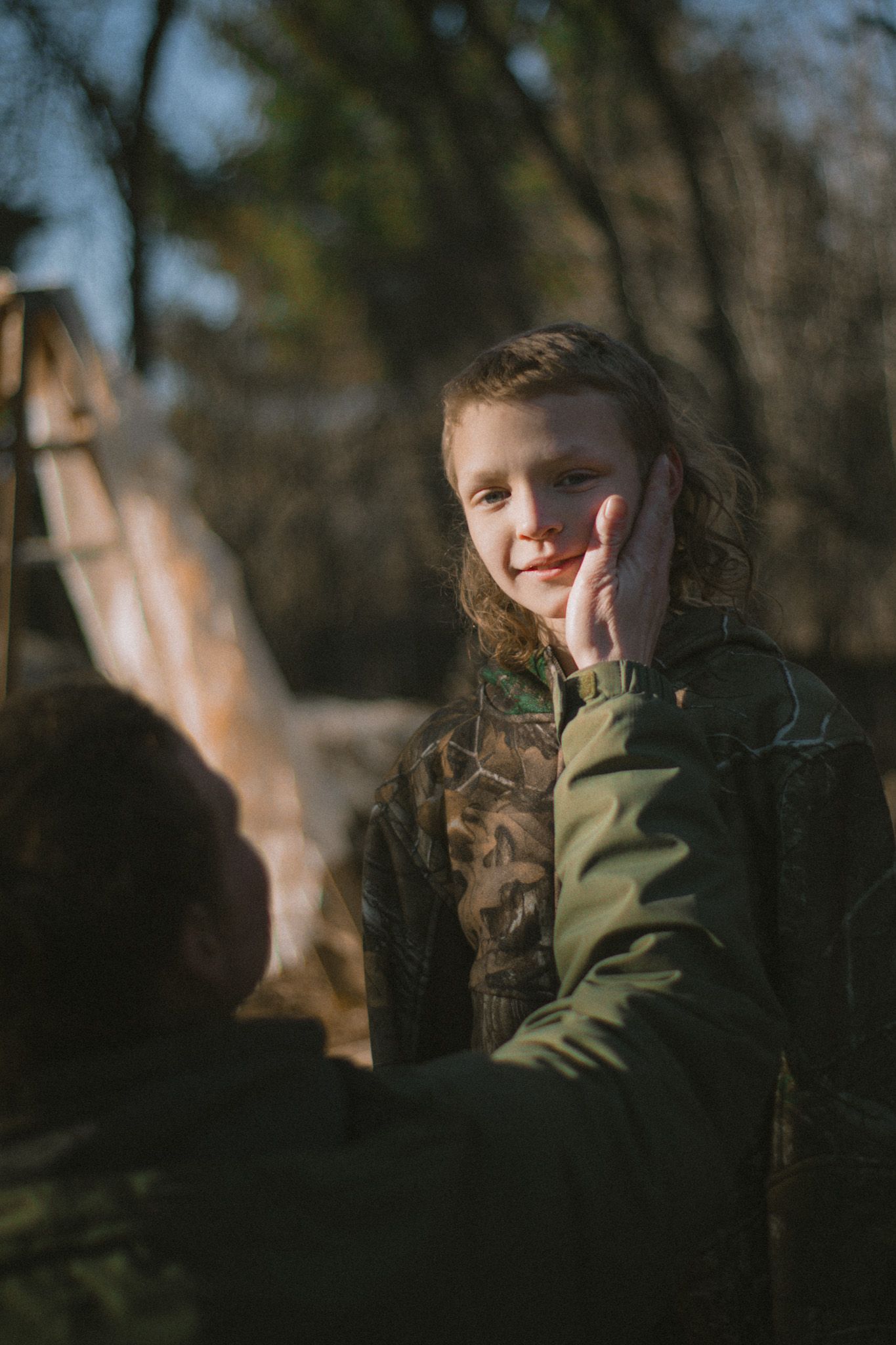 A smiling child in camouflage jacket being gently touched on the cheek outdoors in soft natural light.