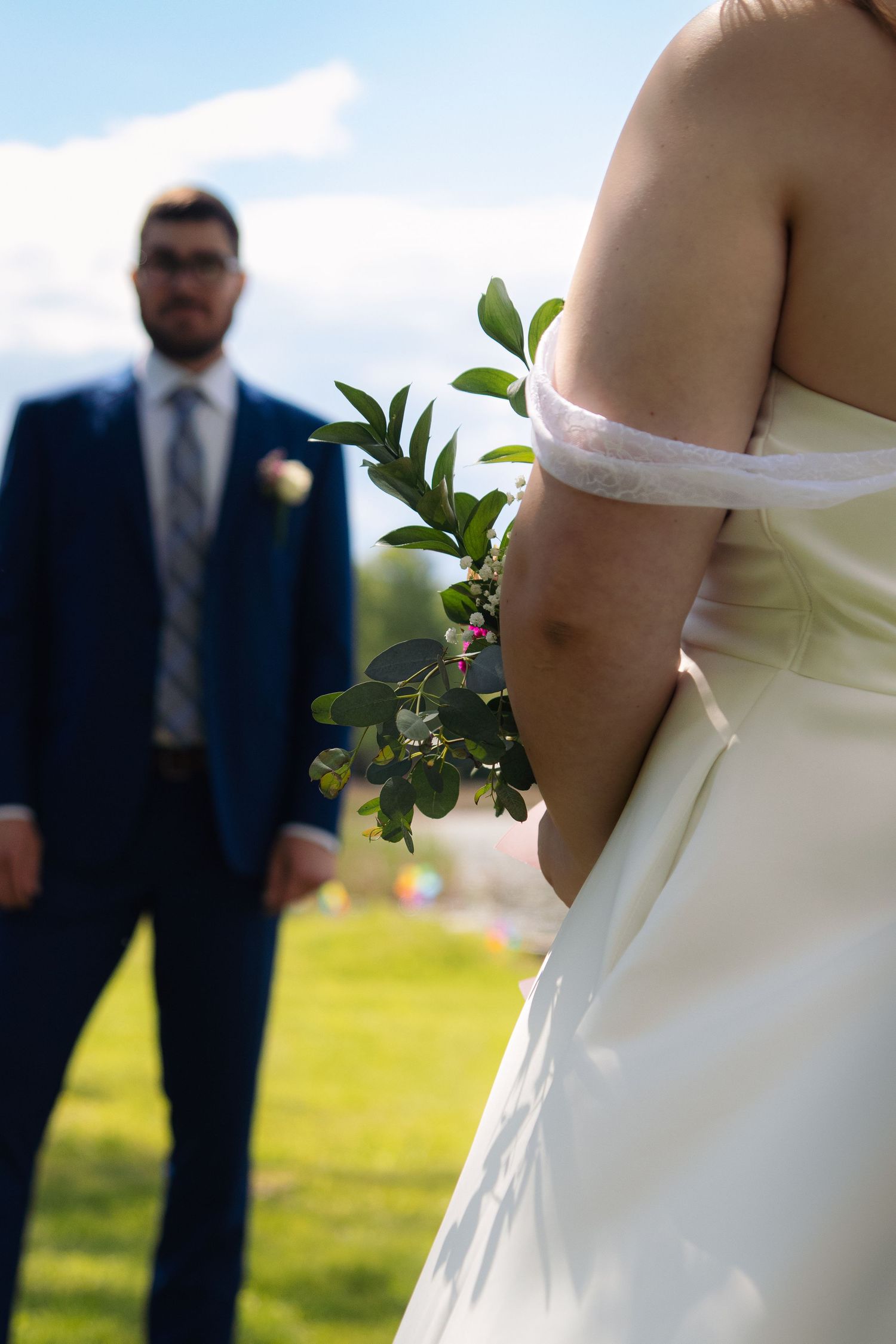 An artistic wedding photo showing a bride's dress detail with the groom standing in the background.