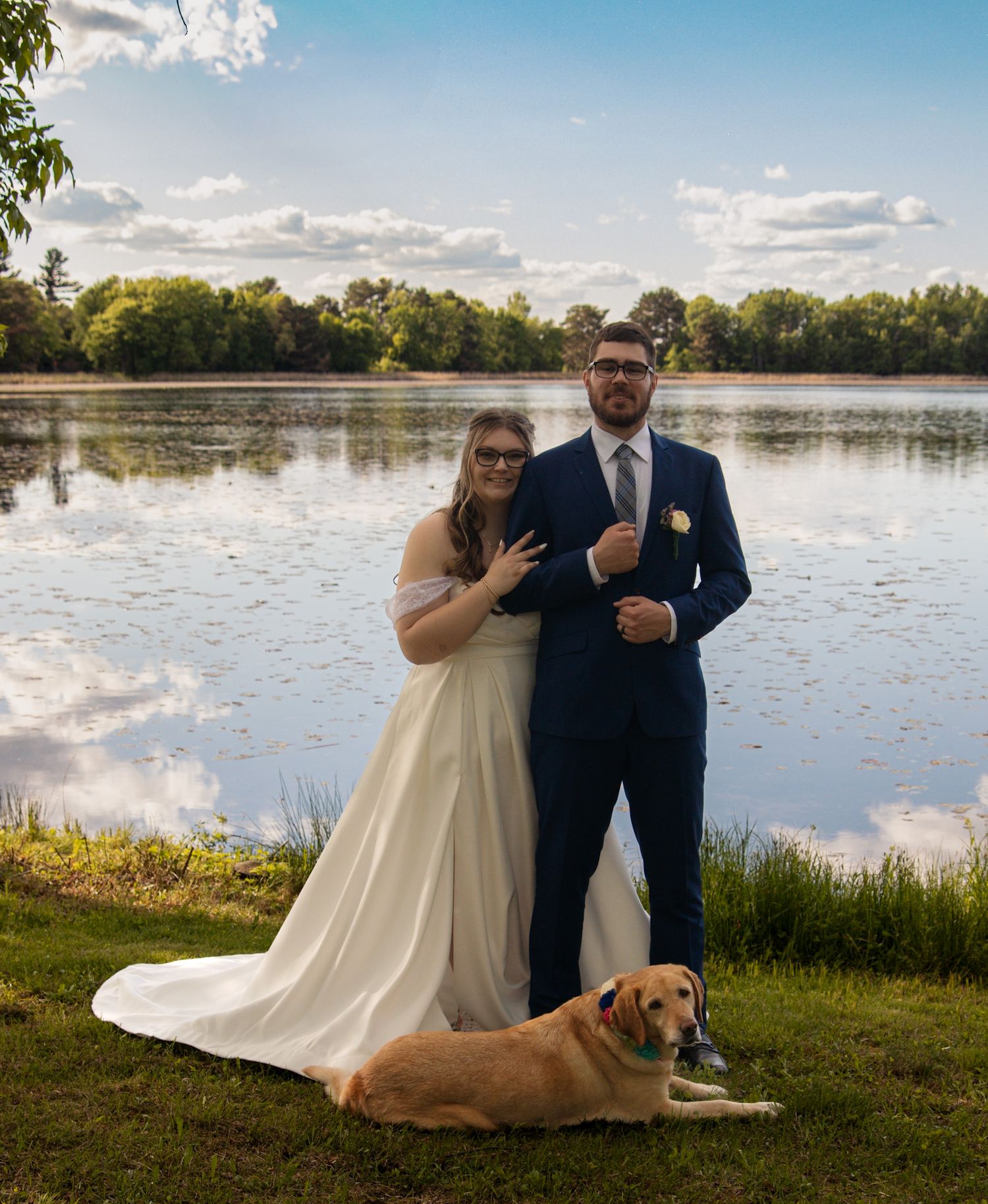 A bride and groom pose with their dog by a serene lake on their wedding day, with cloudy skies and greenery around them.