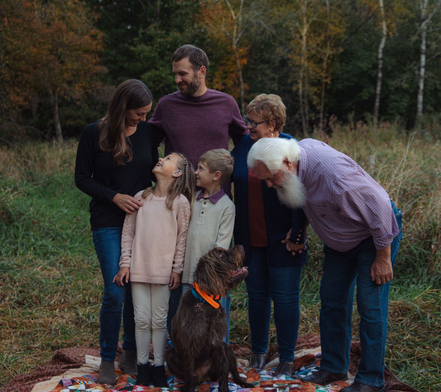 A family and their brown dog gather together outdoors on an autumn day with fall foliage in the background.