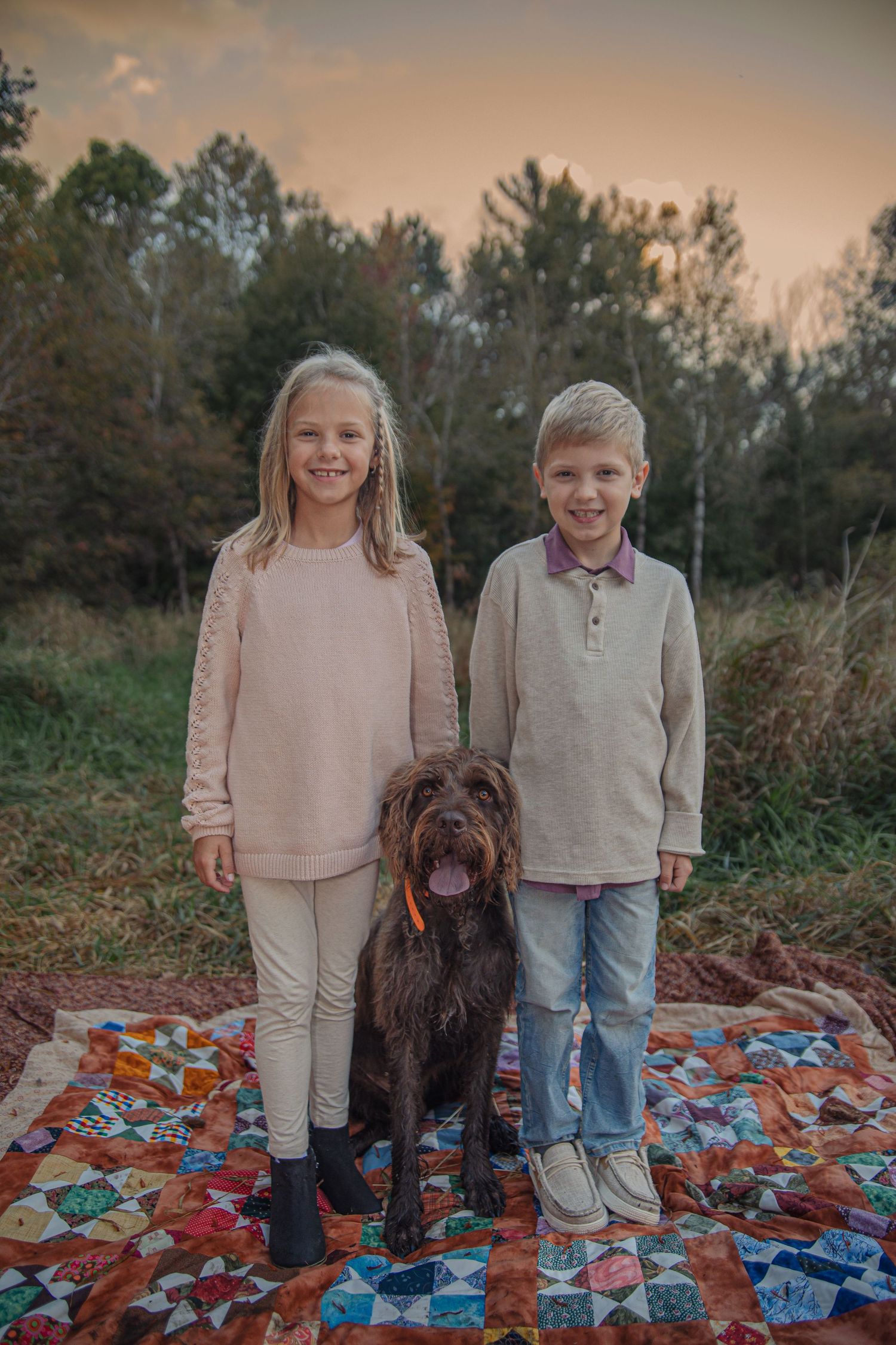 Two children and their brown dog pose together on a colorful patchwork quilt outdoors during sunset.