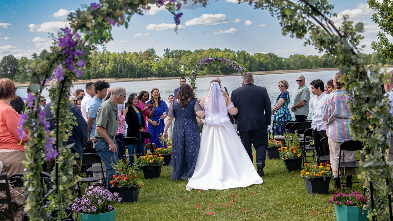 Panoramic view of an outdoor garden wedding ceremony with guests gathered under blue skies and floral decorations.