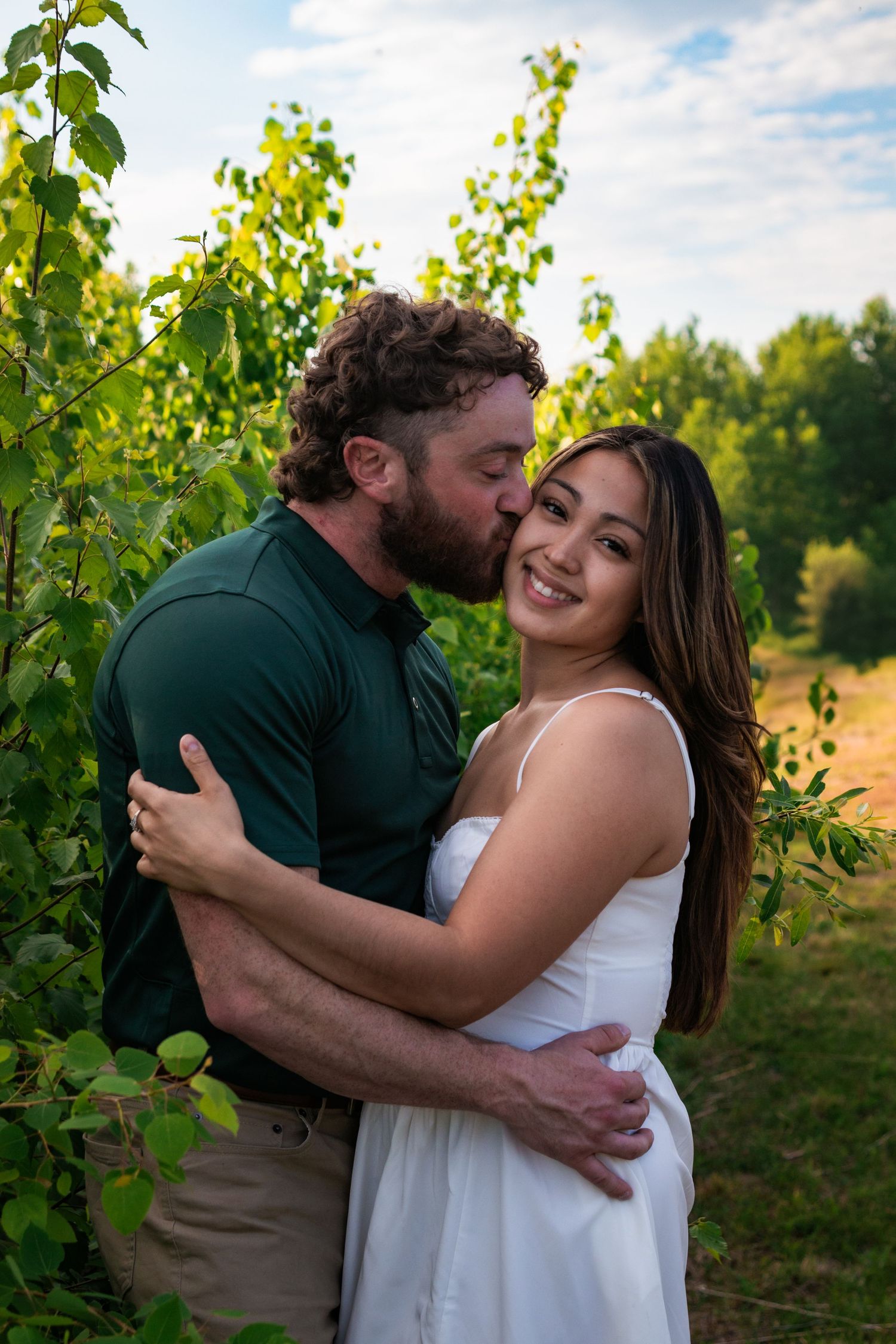A romantic couple embraces tenderly among lush green grapevines at sunset in a vineyard.