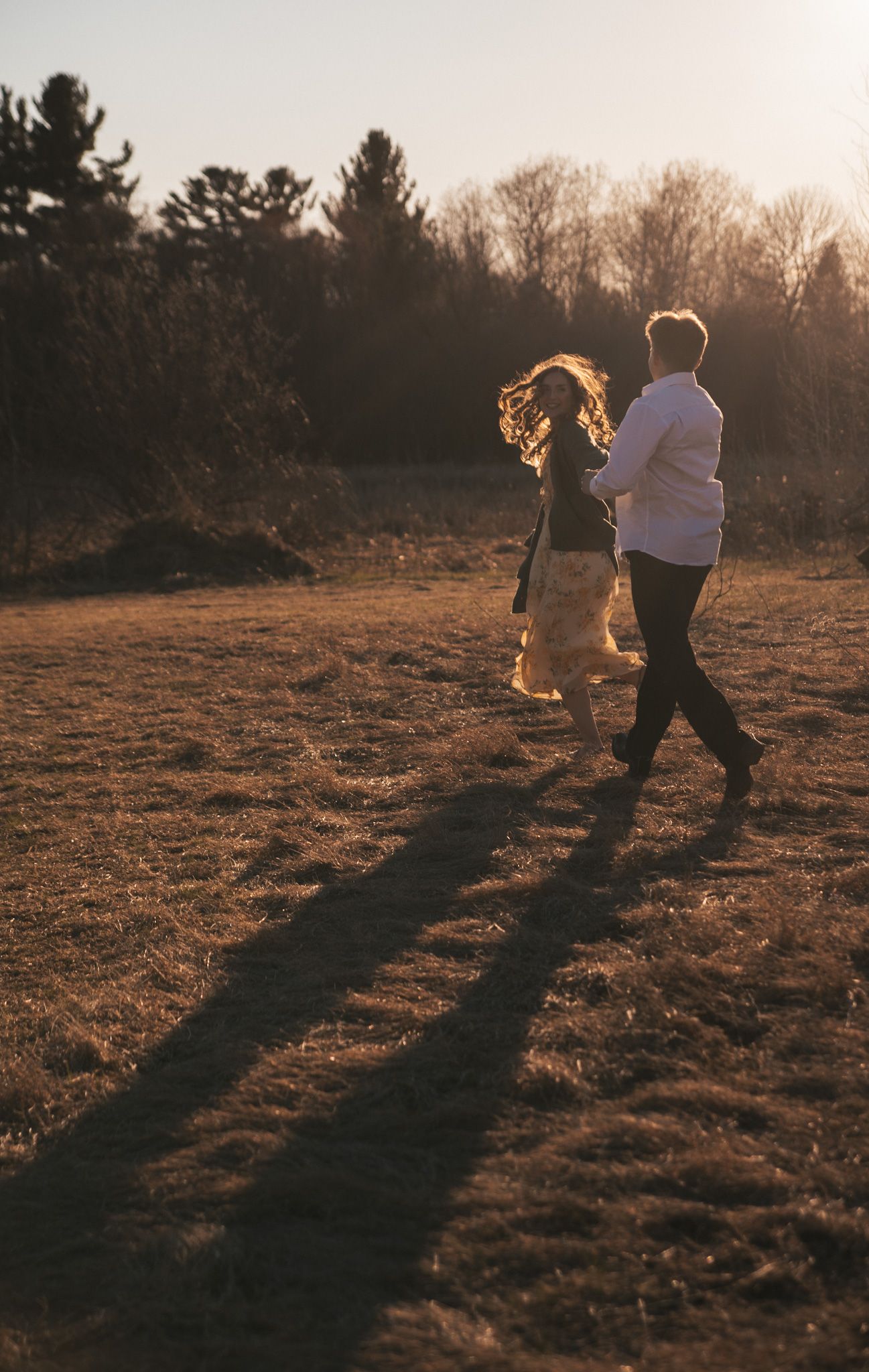 Couple running hand in hand through golden field at sunset with long shadows and trees in background.