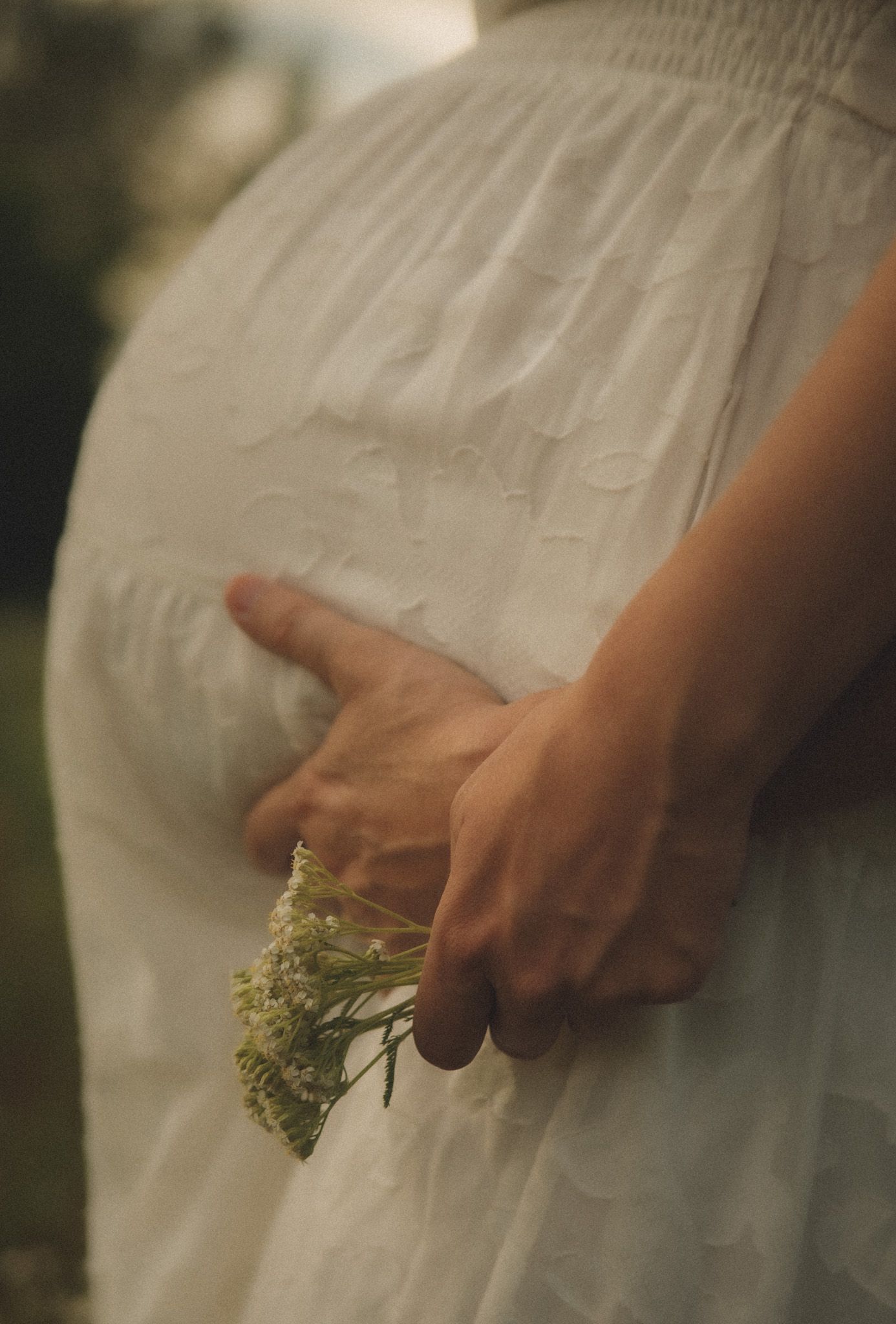 Person in white dress holding small wildflowers behind their back in a soft, dreamy outdoor setting.