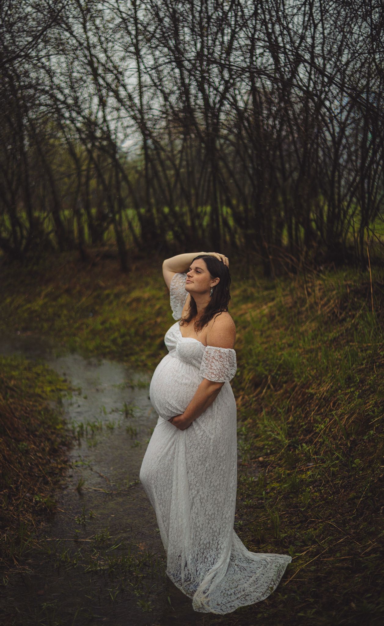 Pregnant woman in white lace off-shoulder gown poses outdoors near a stream surrounded by bare trees.
