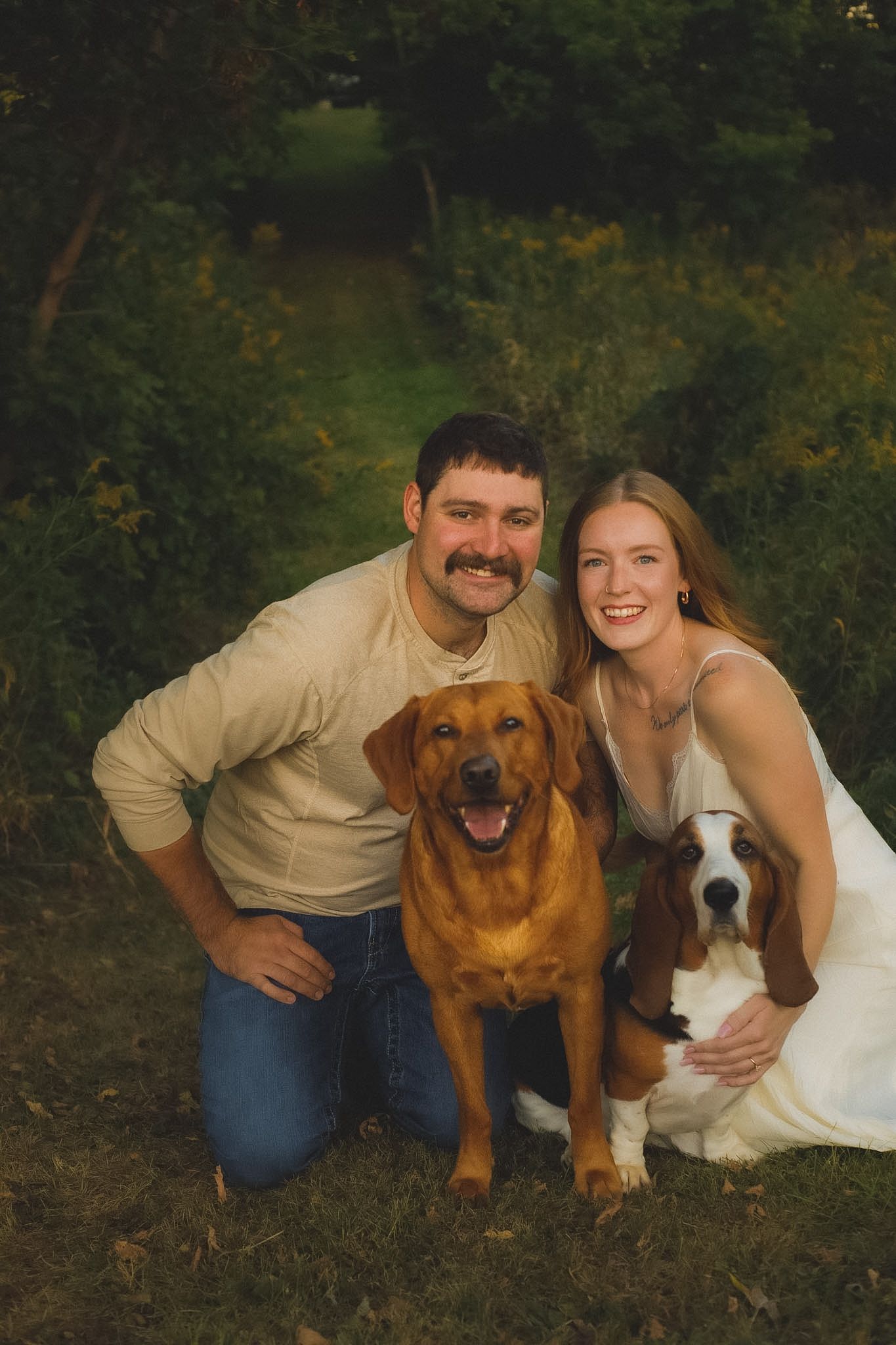 Smiling couple kneeling outdoors with a golden Labrador and Basset Hound in a lush green forest setting.