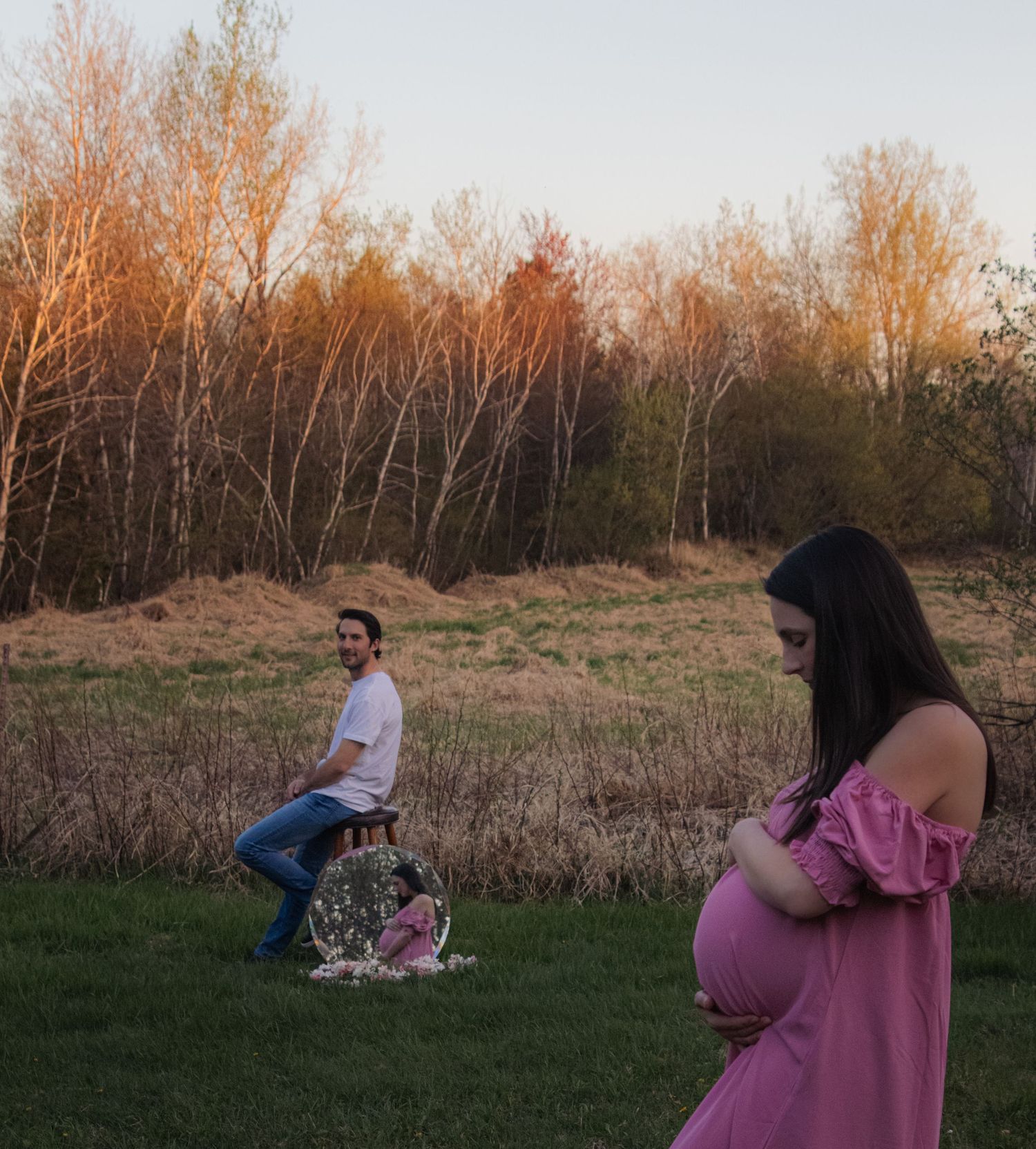 A couple sits together in a grassy field at sunset while trees glow with warm golden light in the background.