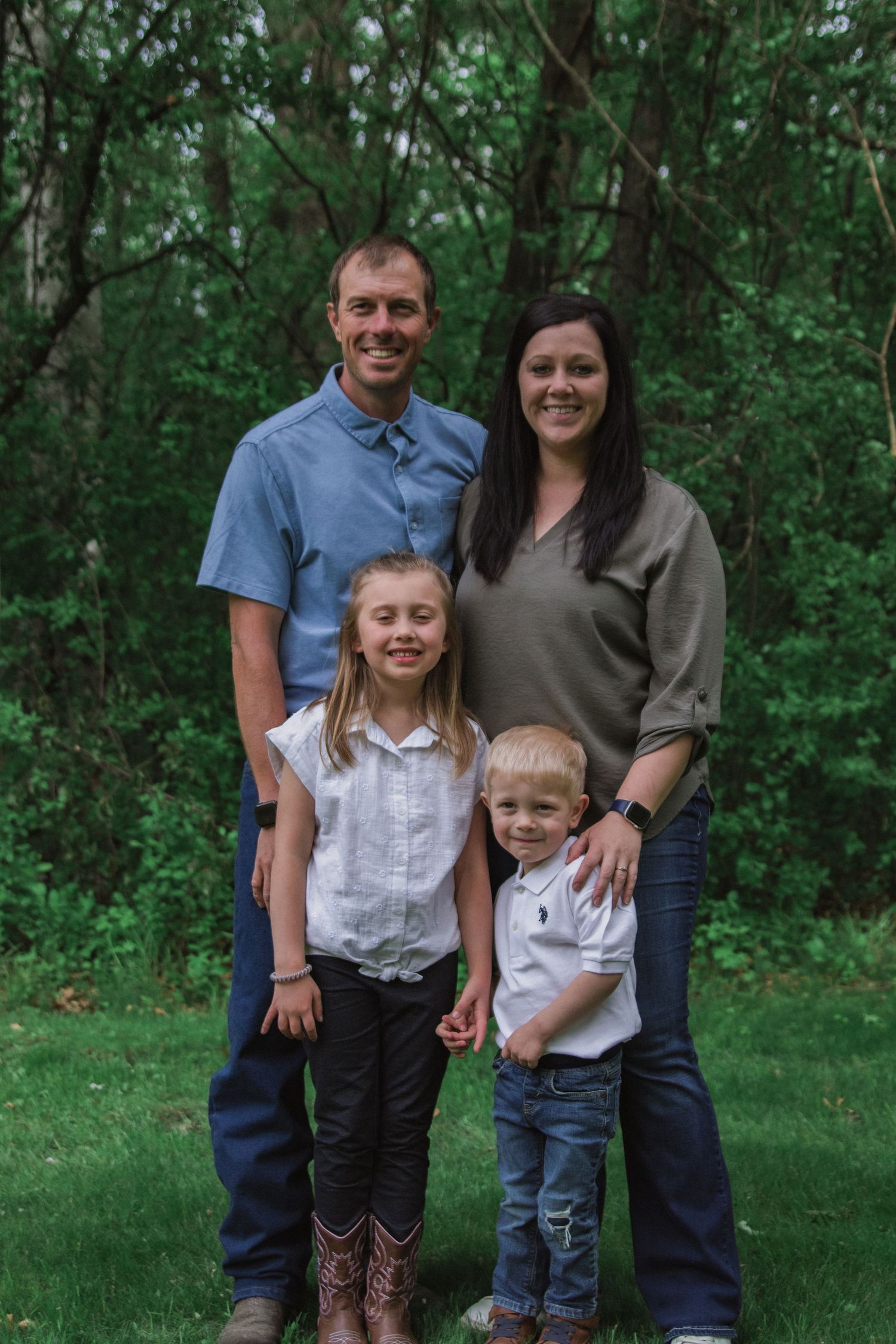 A couple with two children pose for a portrait outdoors against a green wooded background.