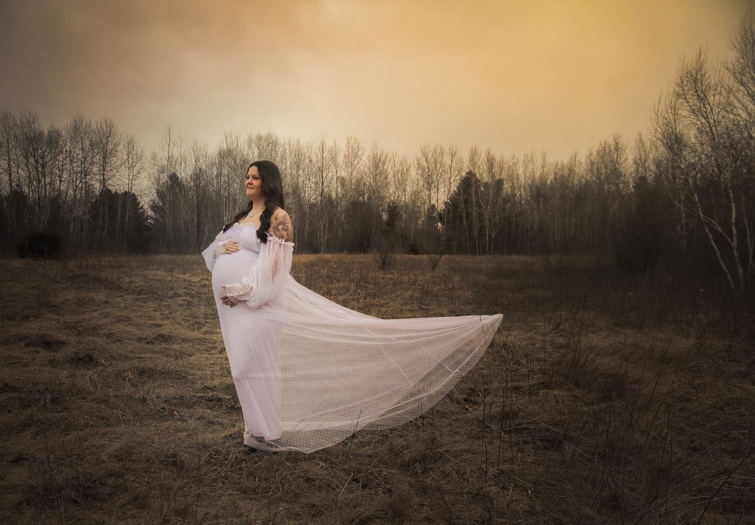 Pregnant woman in flowing white lace gown standing in golden field at sunset.