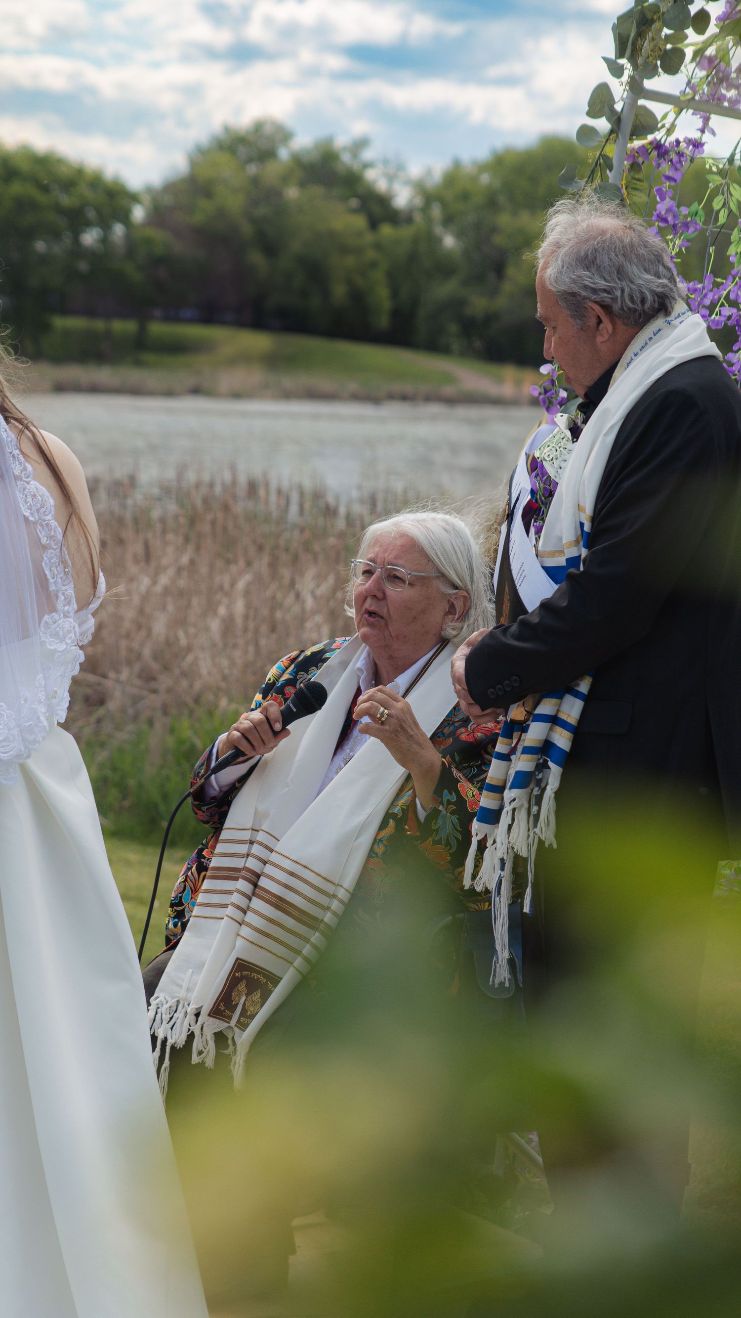 Jewish wedding ceremony with people wearing traditional prayer shawls outdoors against a natural landscape.