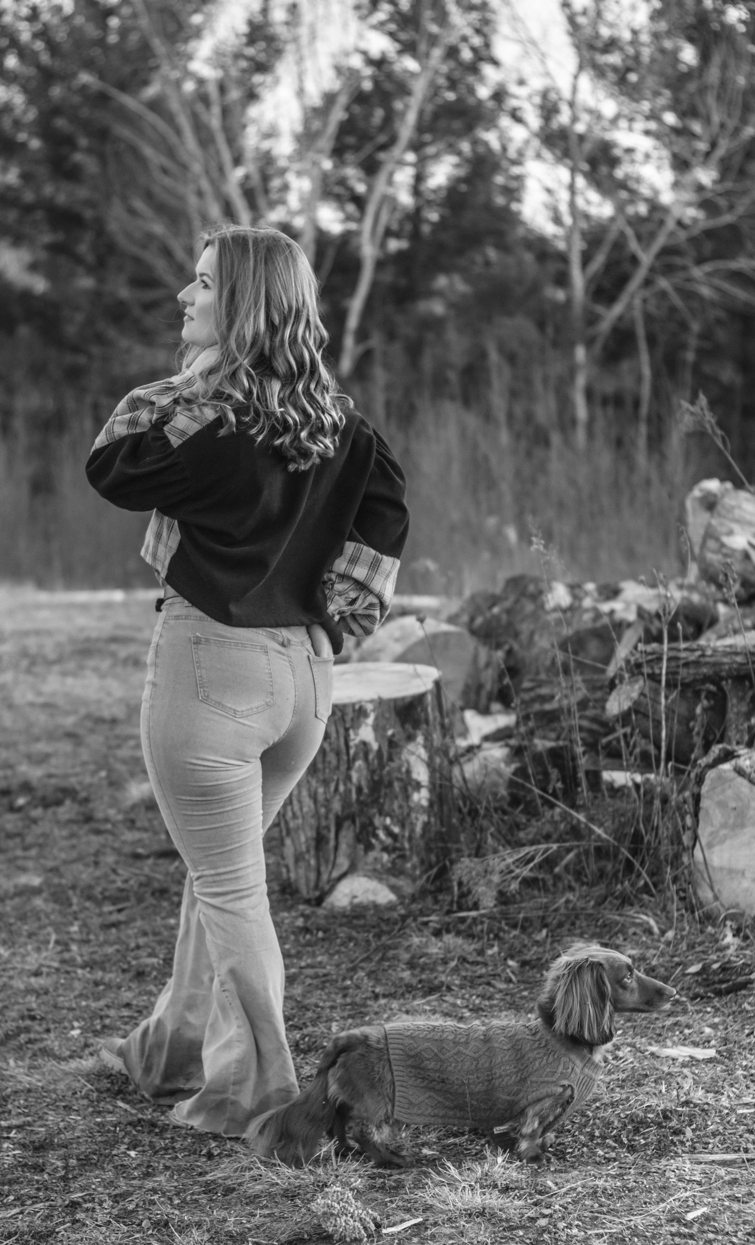 Black and white photo of a woman standing outdoors with a dachshund dog near a tree stump in a wooded area.