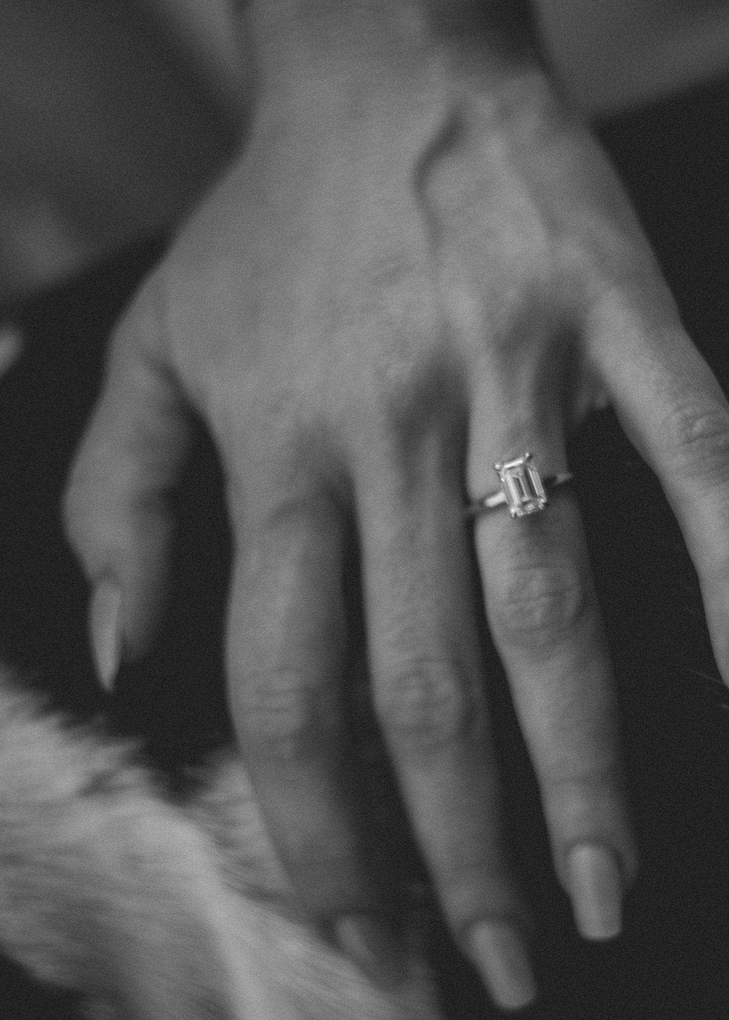 Black and white close-up of a hand wearing an elegant emerald-cut diamond solitaire engagement ring.