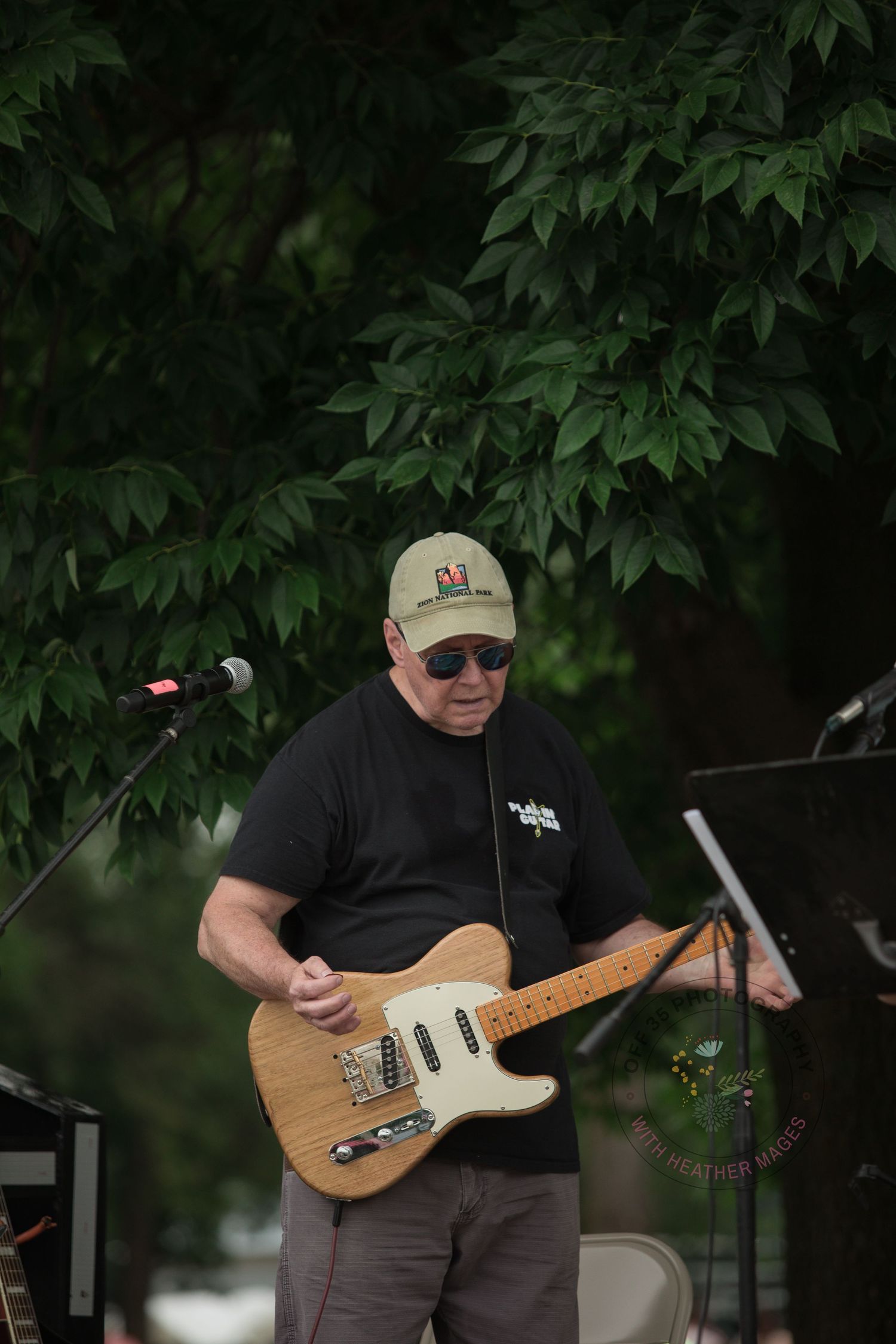 A musician performs outdoors with a natural maple Telecaster-style electric guitar against a leafy background.
