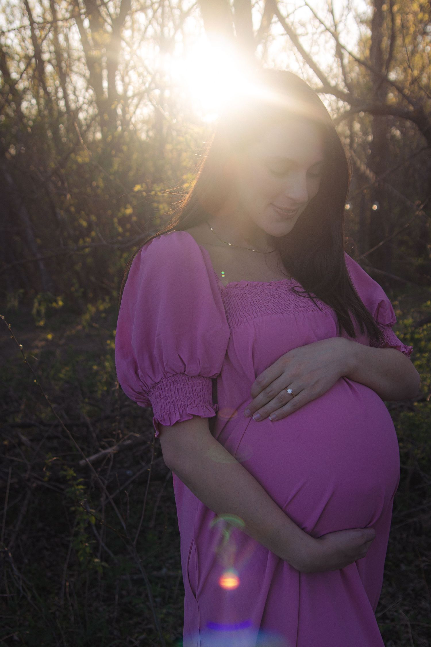 A person in a purple maternity dress stands in a sunlit forest during a photoshoot at golden hour.