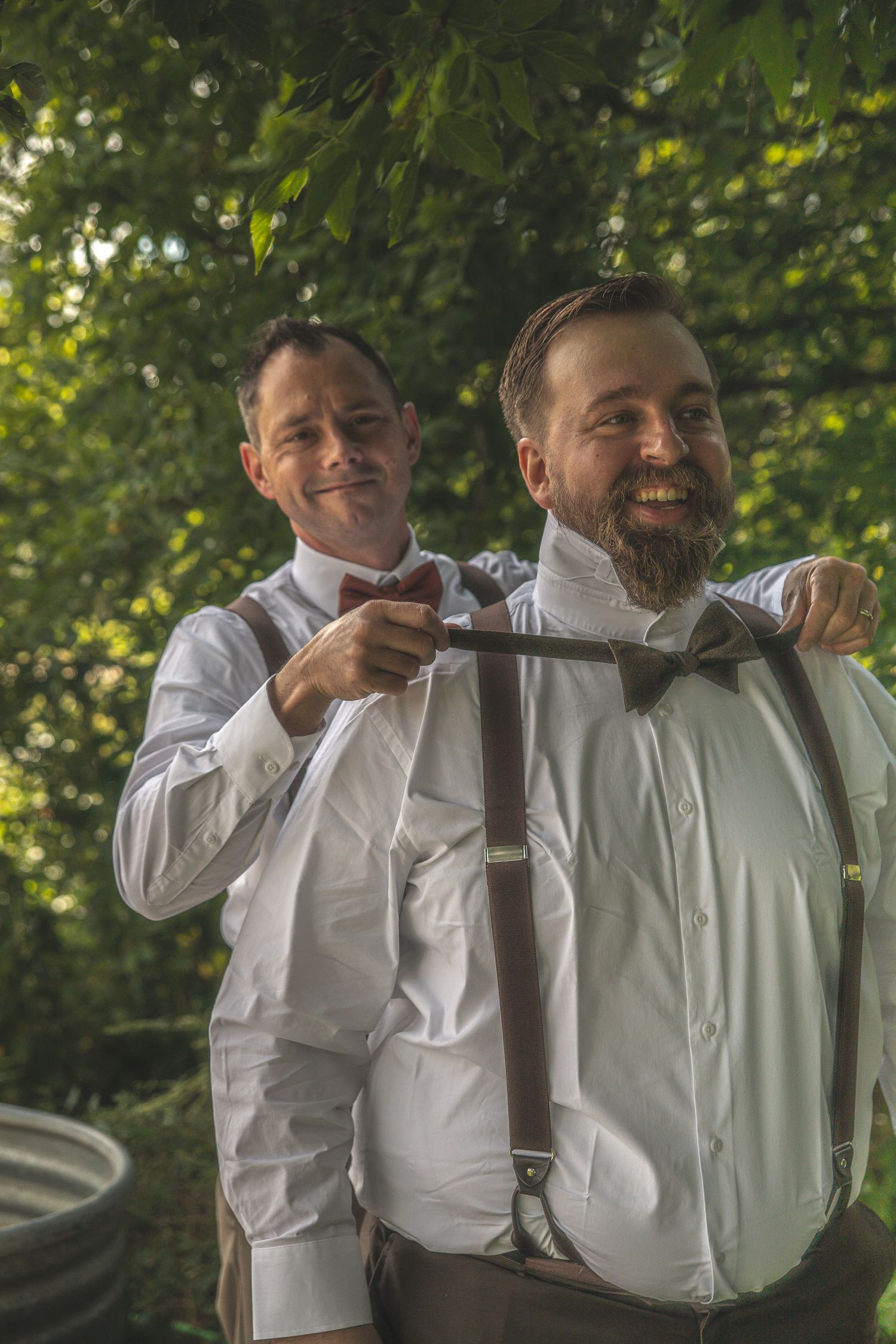 Two men in white dress shirts and suspenders share a laugh together outdoors with green foliage in the background.