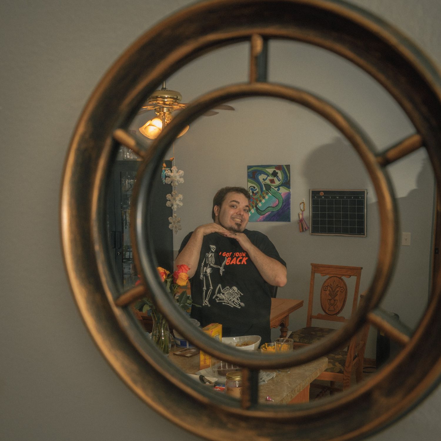 A man takes a selfie in a decorative round bronze mirror reflecting his room with posters and a ceiling fan.